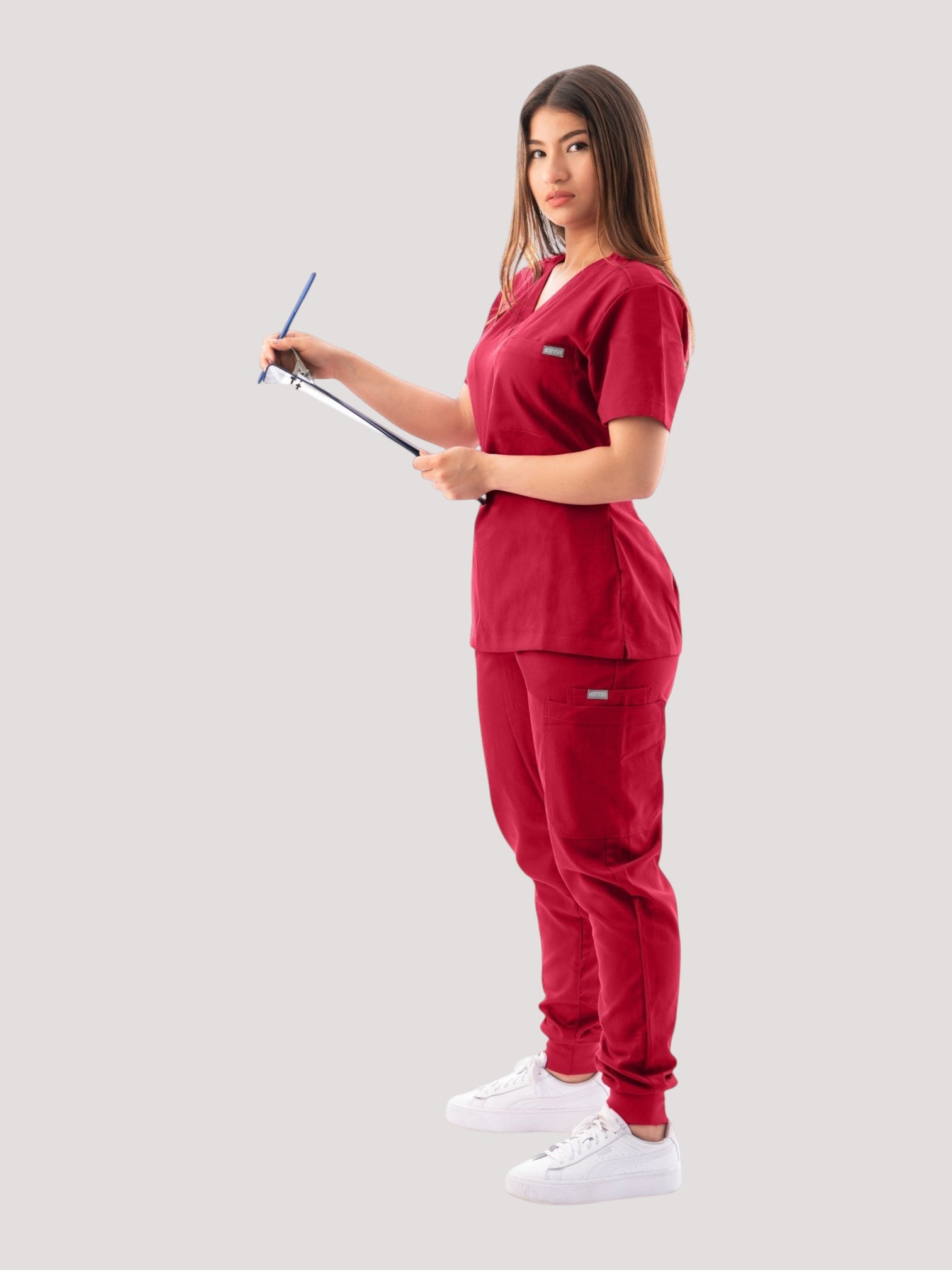 Kotyss women's burgundy V-neck scrub top and jogger pants set worn by a model holding a clipboard.