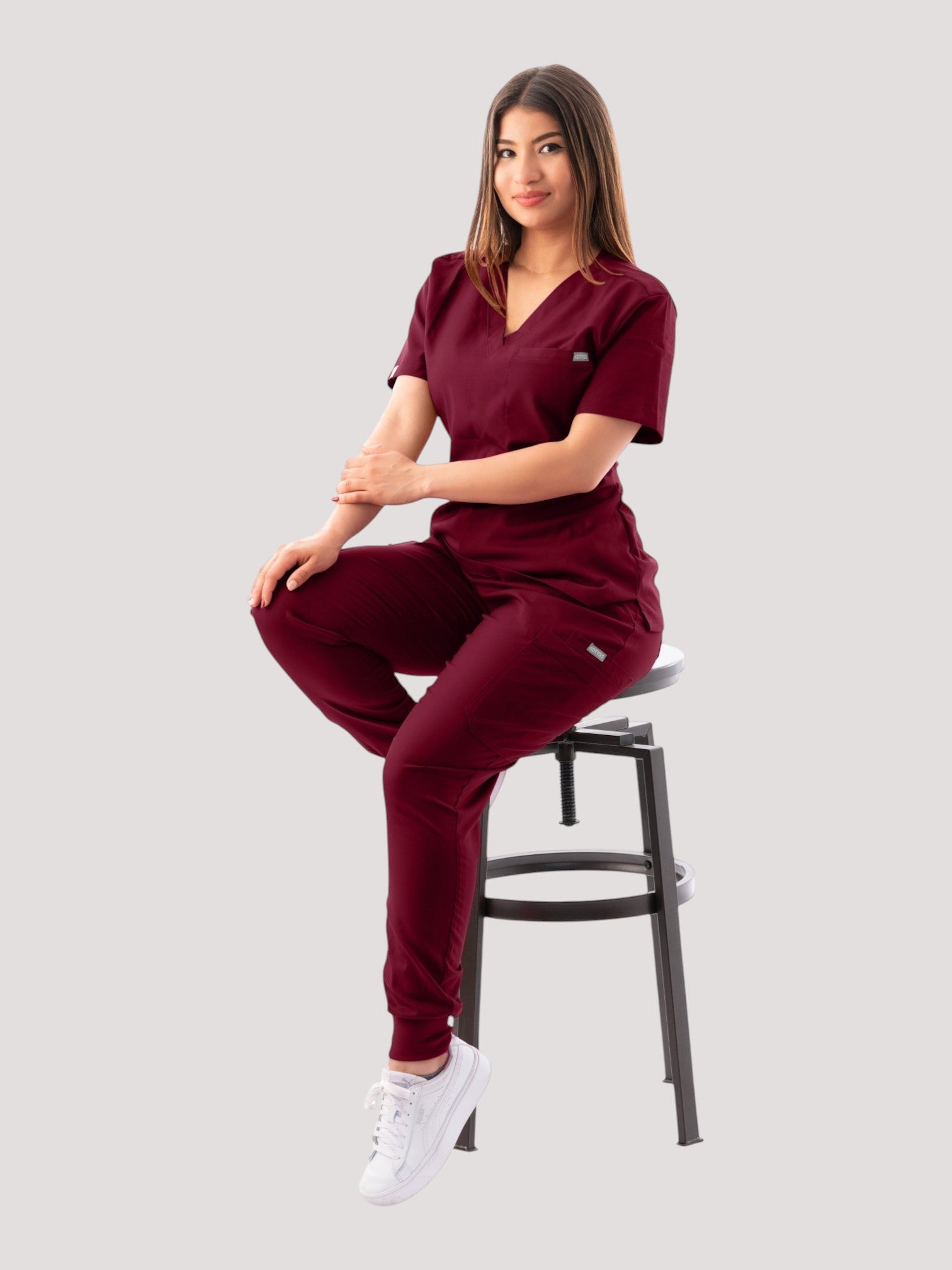 A woman wearing a burgundy Kotyss V-neck scrub top and matching jogger pants while sitting on a stool.