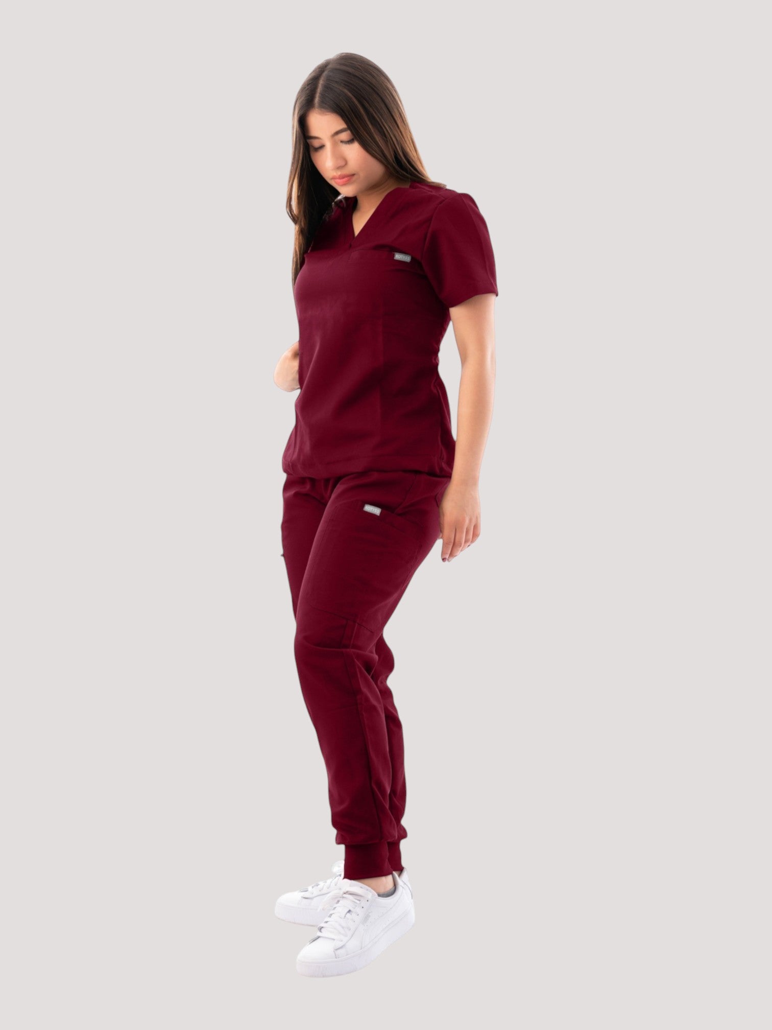 A woman wearing a burgundy Kotyss V-neck scrub top and matching jogger pants with multiple pockets.
