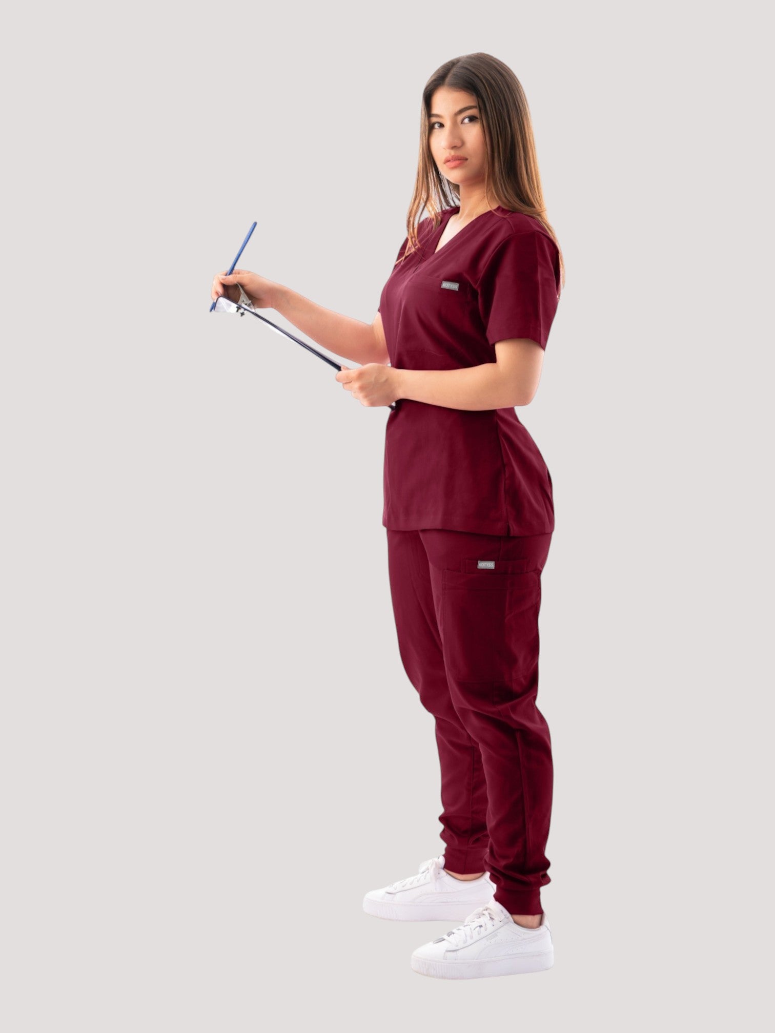 A woman wearing a burgundy Kotyss V-neck scrub top and matching jogger pants while holding a clipboard.