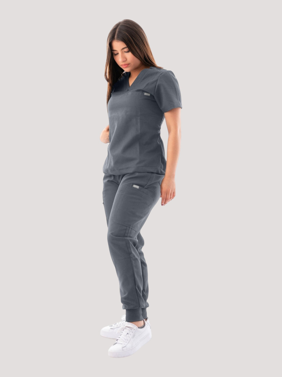 Model wearing a charcoal grey Kotyss V-neck scrub top and matching multi-pocket jogger pants with white sneakers.