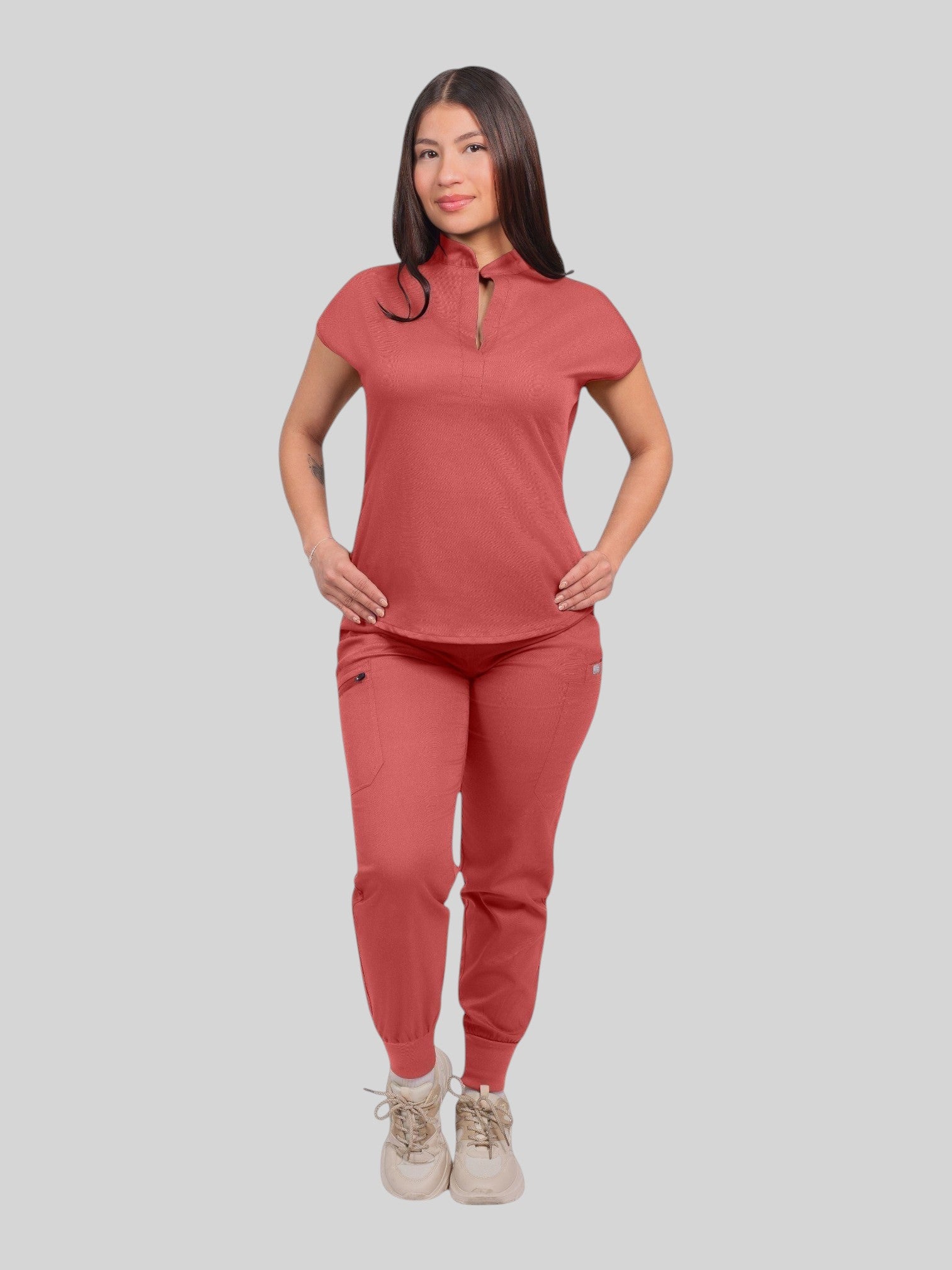 A woman wearing a Kotyss coral collared scrub top and matching jogger pants with multiple pockets.