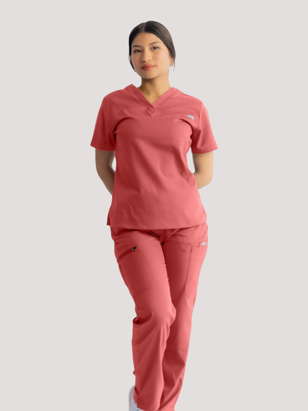 A woman wearing Kotyss coral pink straight cut scrub pants and matching v-neck top with multiple utility pockets.