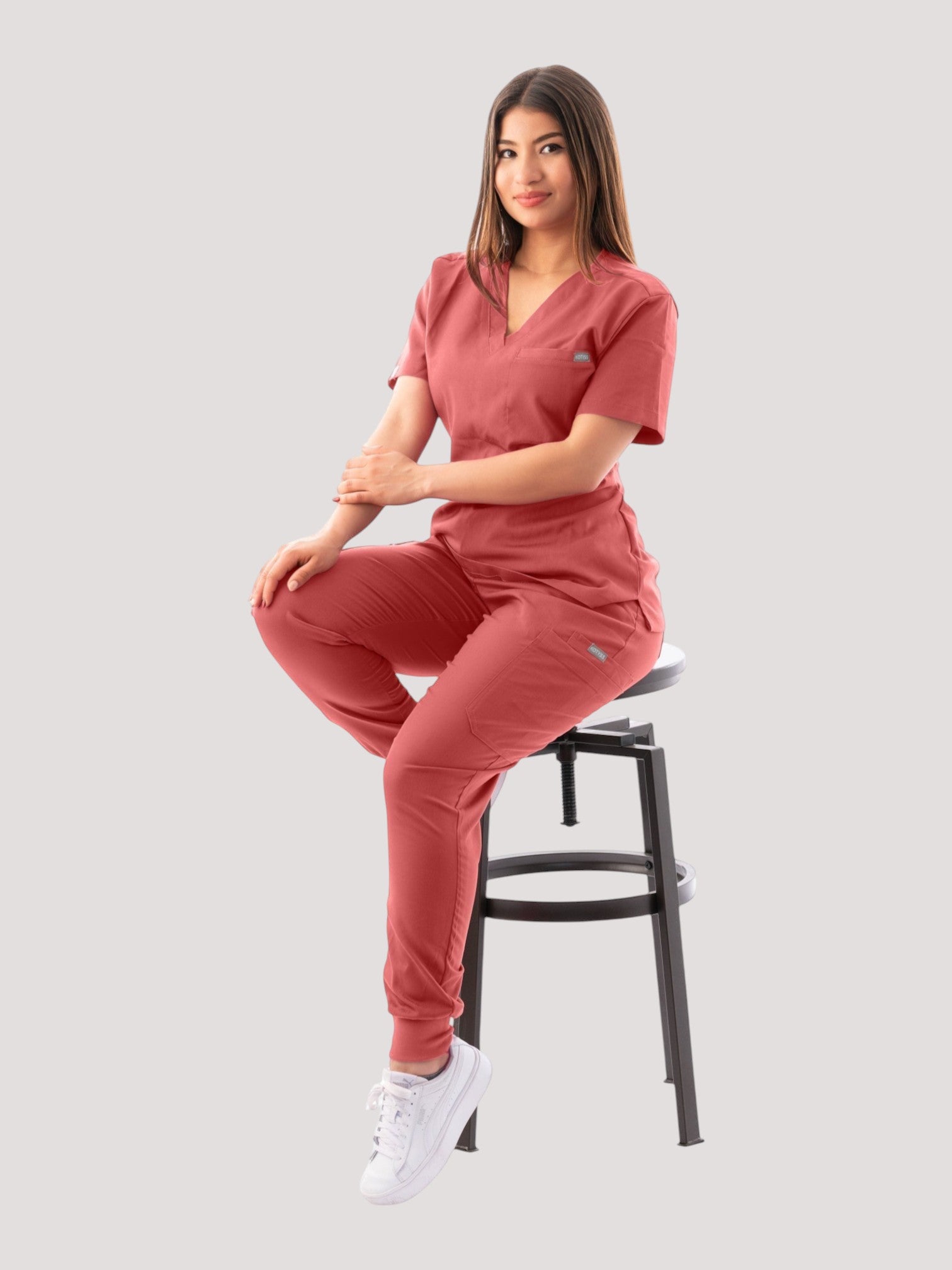 A woman wearing a Kotyss coral V-neck scrub top and matching jogger pants, sitting on a stool against a white background.
