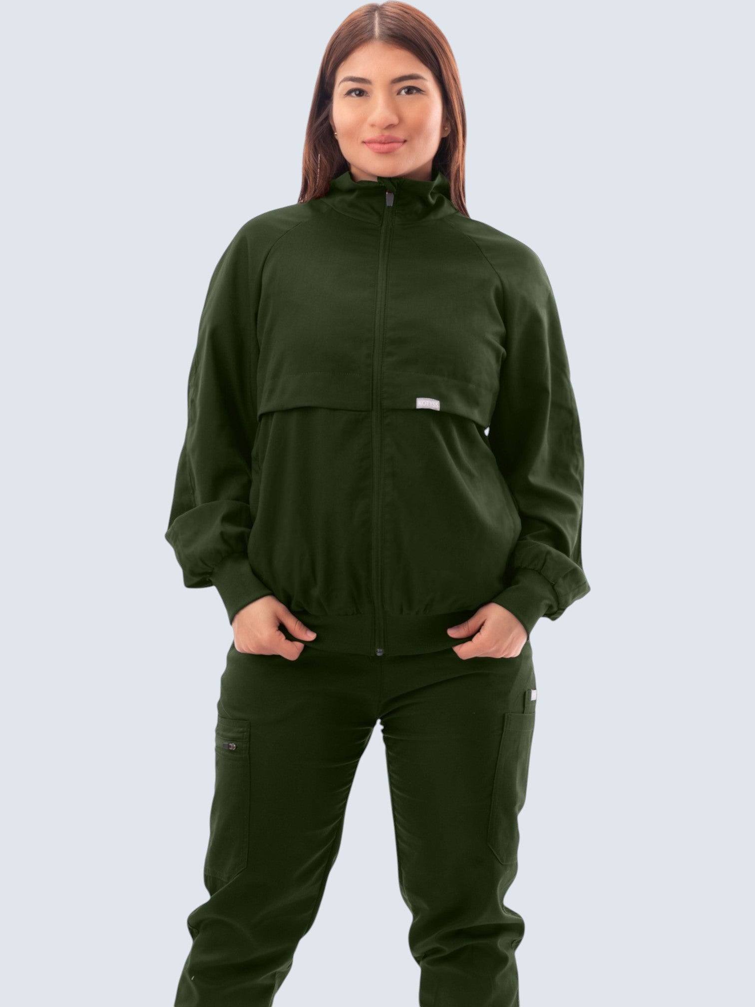 A woman wearing a dark green Kotyss scrub jacket with a high windbreaker neck, ribbed cuffs, and a tailored fit.