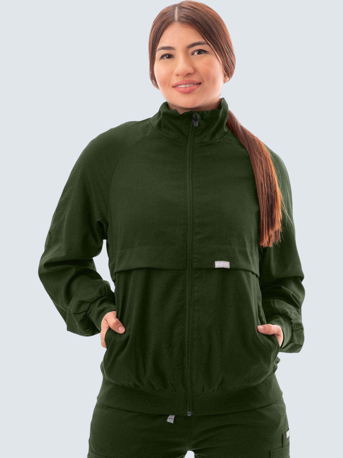 A woman wearing a dark green Kotyss scrub jacket with a high neck, zip front, and ribbed cuffs against a white background