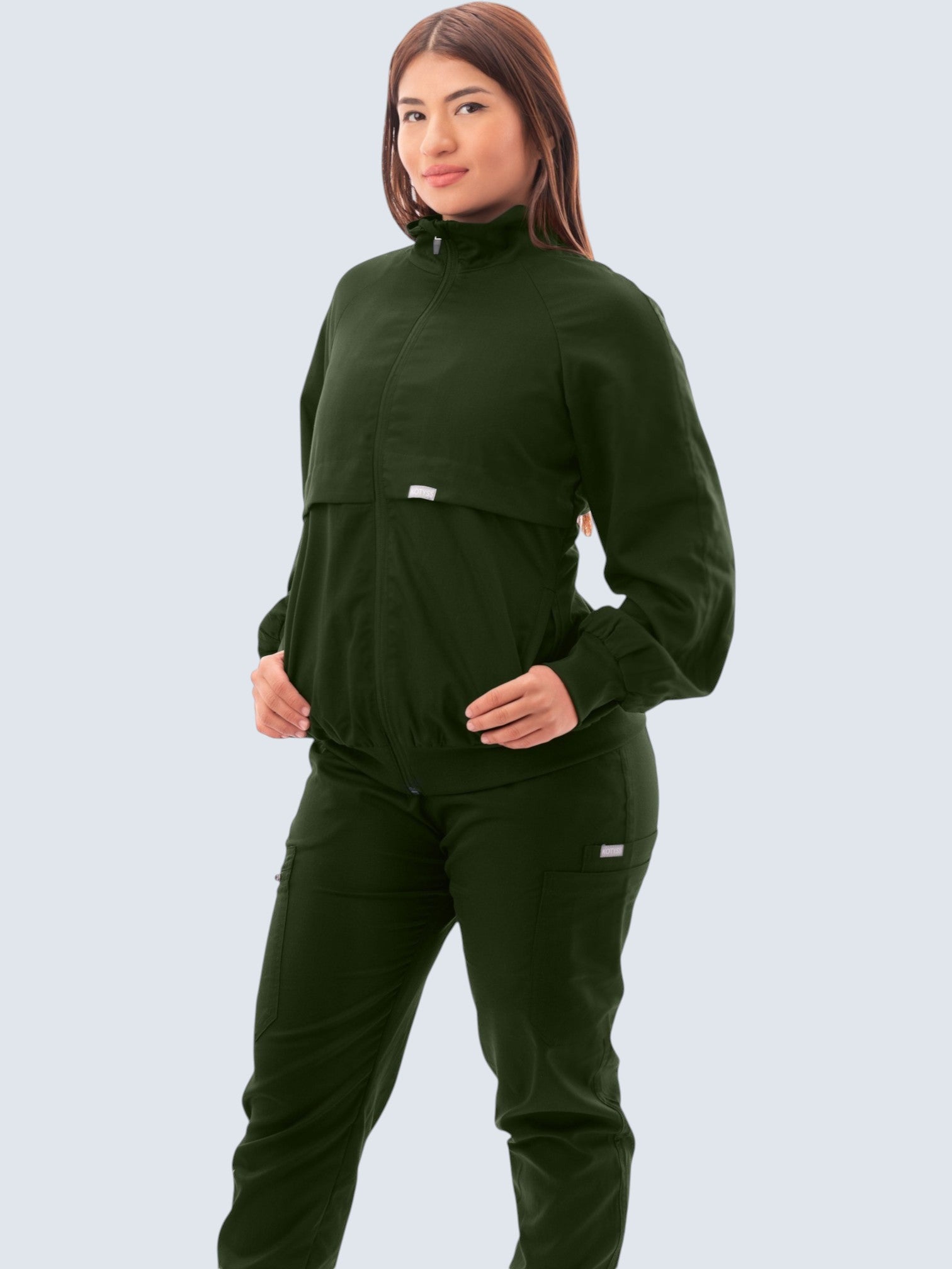 A woman wearing a dark green Kotyss scrub jacket with a high windbreaker neck, ribbed cuffs, and functional pockets.