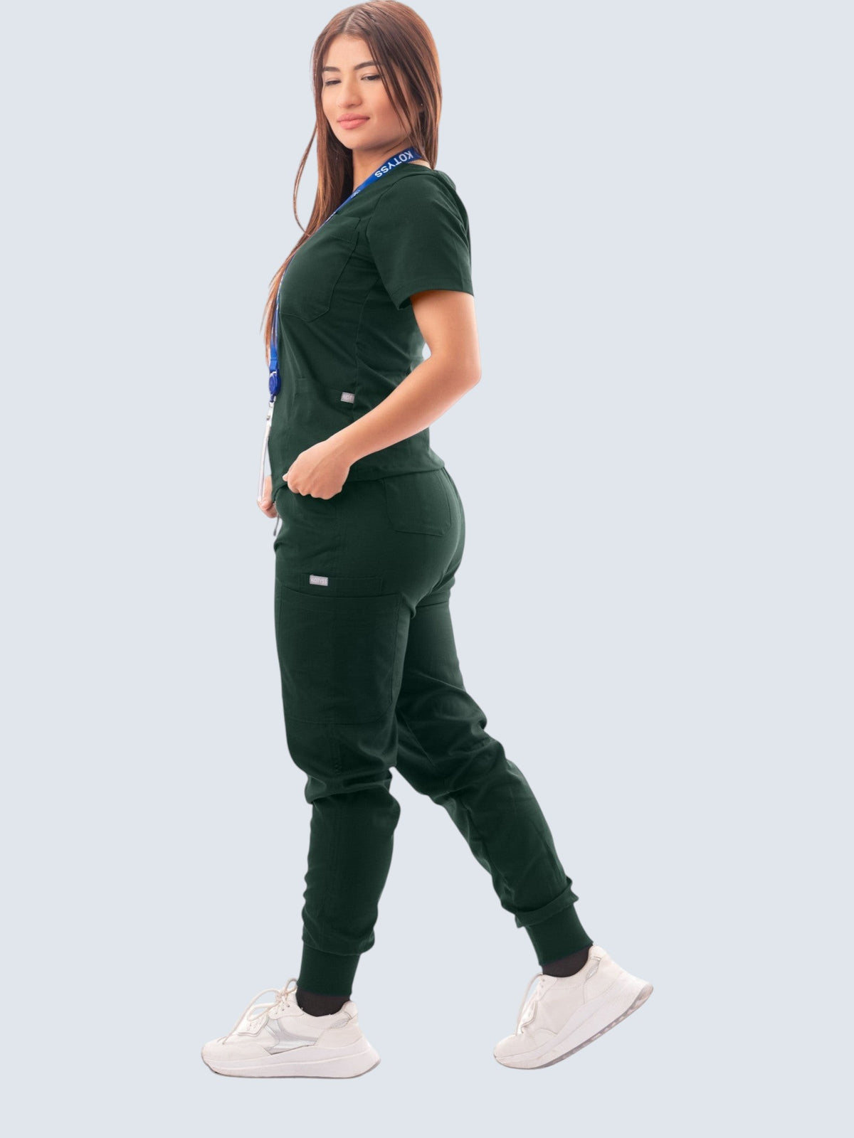Woman wearing a dark green Kotyss V-neck scrub top and multi-pocket jogger pants set with white sneakers.