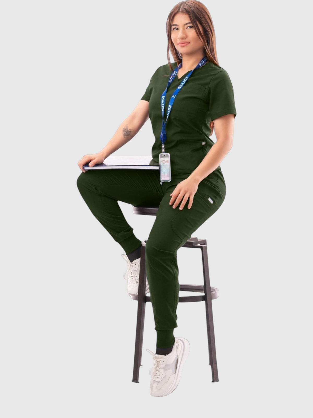 Woman wearing a dark green Kotyss V-neck scrub top and jogger pants set with multiple pockets, sitting on a stool.