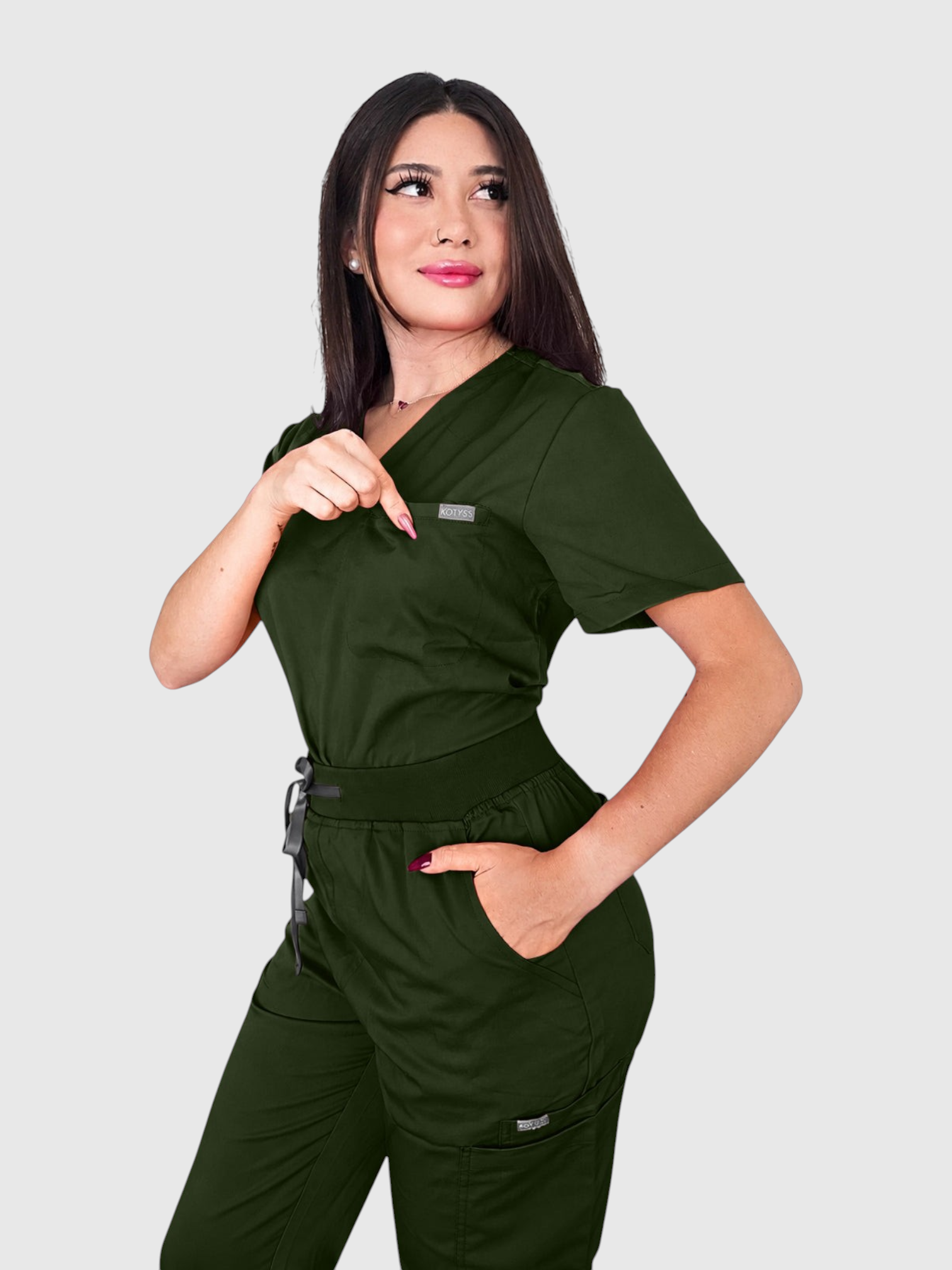 A woman wearing a dark green Kotyss V-neck scrub top and matching jogger pants with multiple pockets.