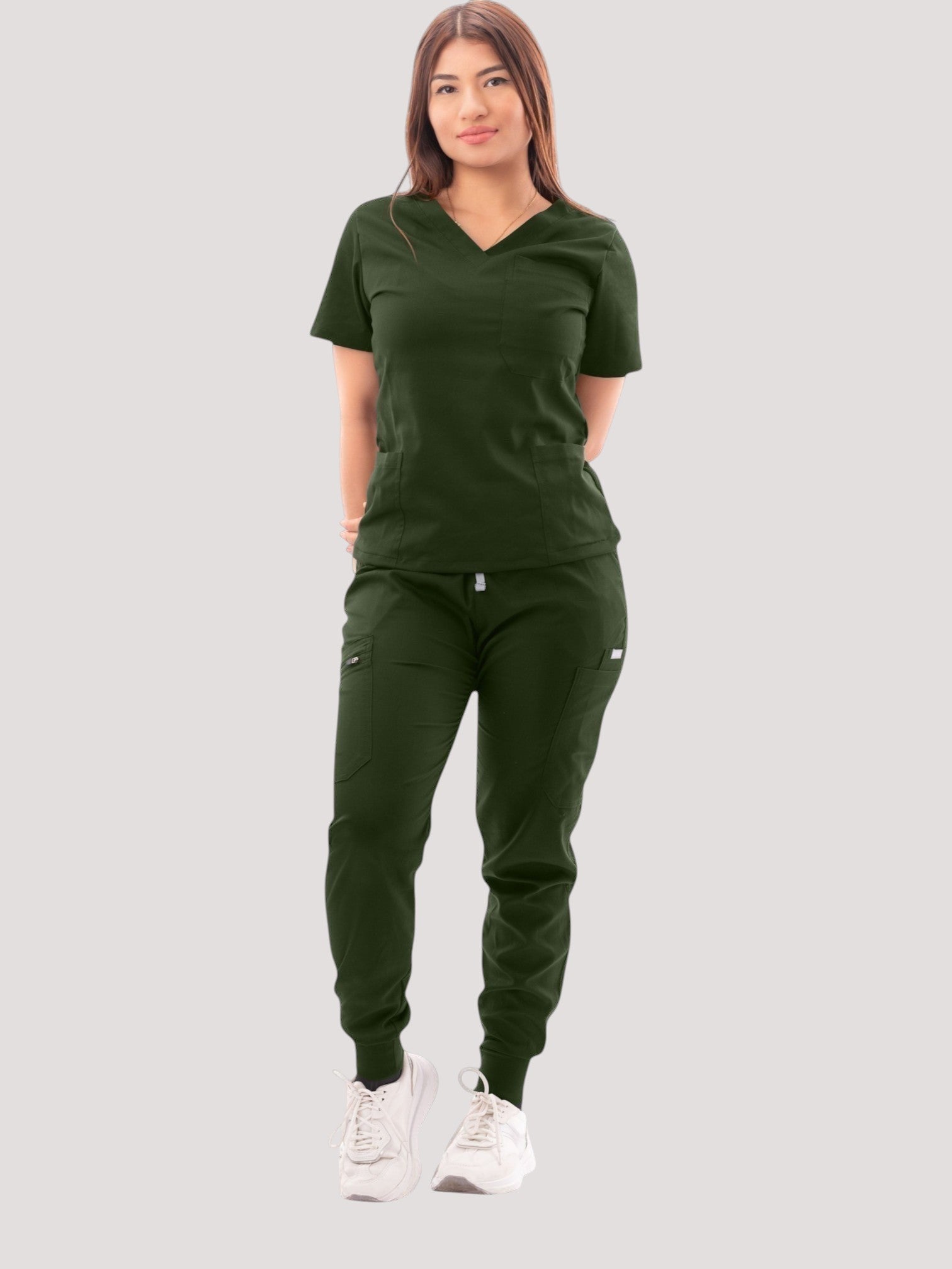 A woman wearing a dark green Kotyss V-neck scrub top and matching multi-pocket jogger pants with white sneakers.