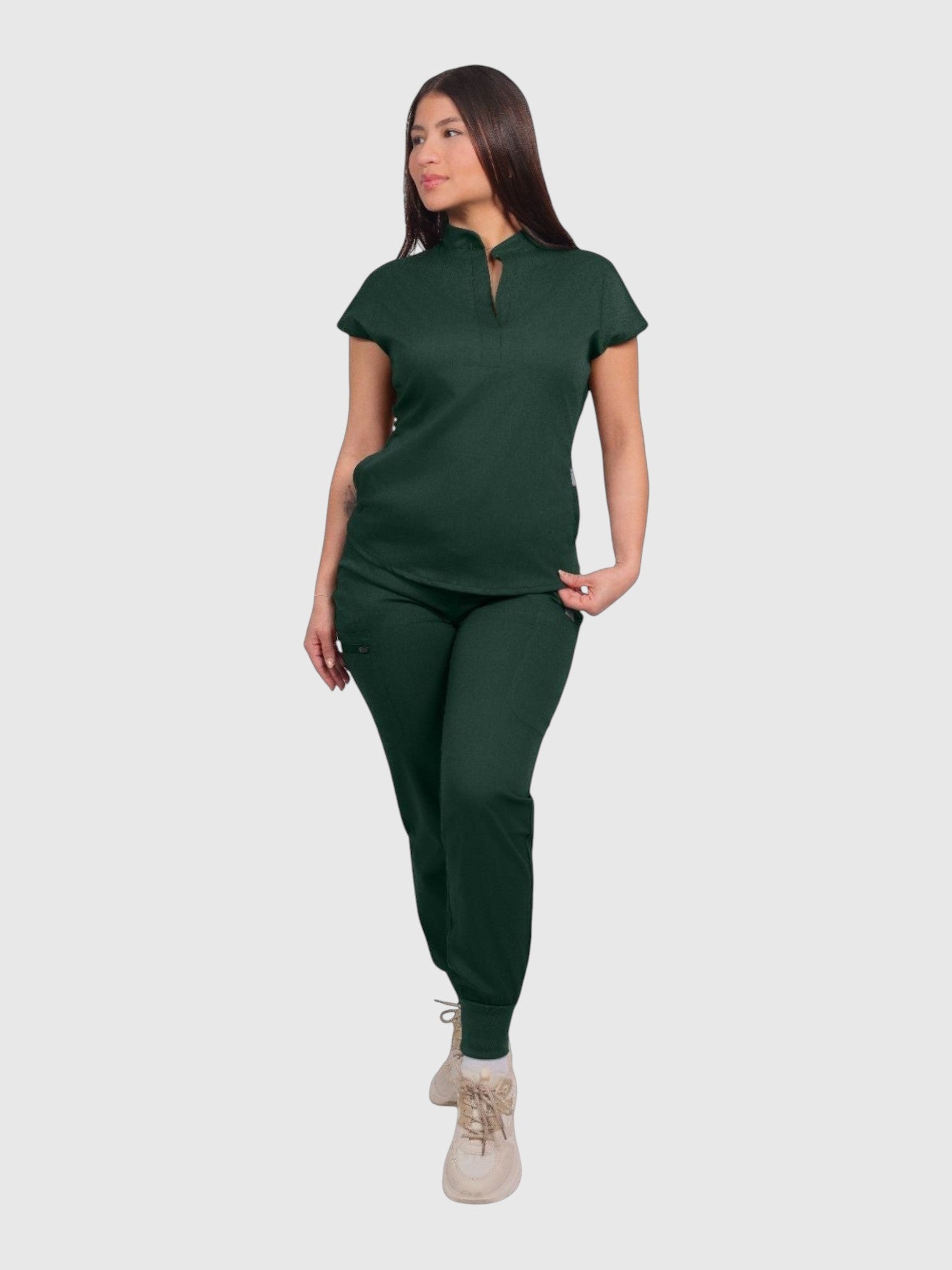 A woman wearing a dark green Kotyss collared scrub top and matching jogger pants with multiple pockets.