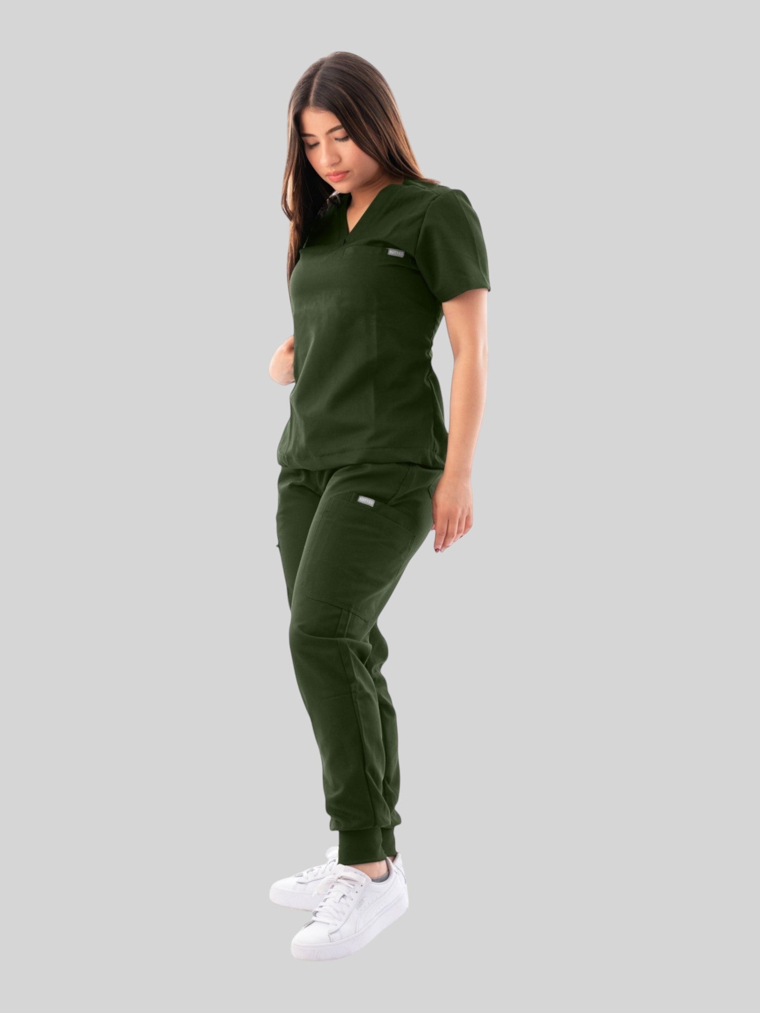 A woman wearing a dark green Kotyss V-neck scrub top and matching jogger pants with multiple pockets.