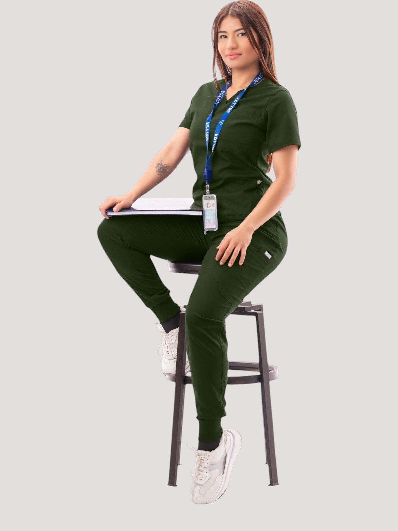 Kotyss women's dark green V-neck scrub top and jogger pants set with multiple pockets on a model sitting on a stool.