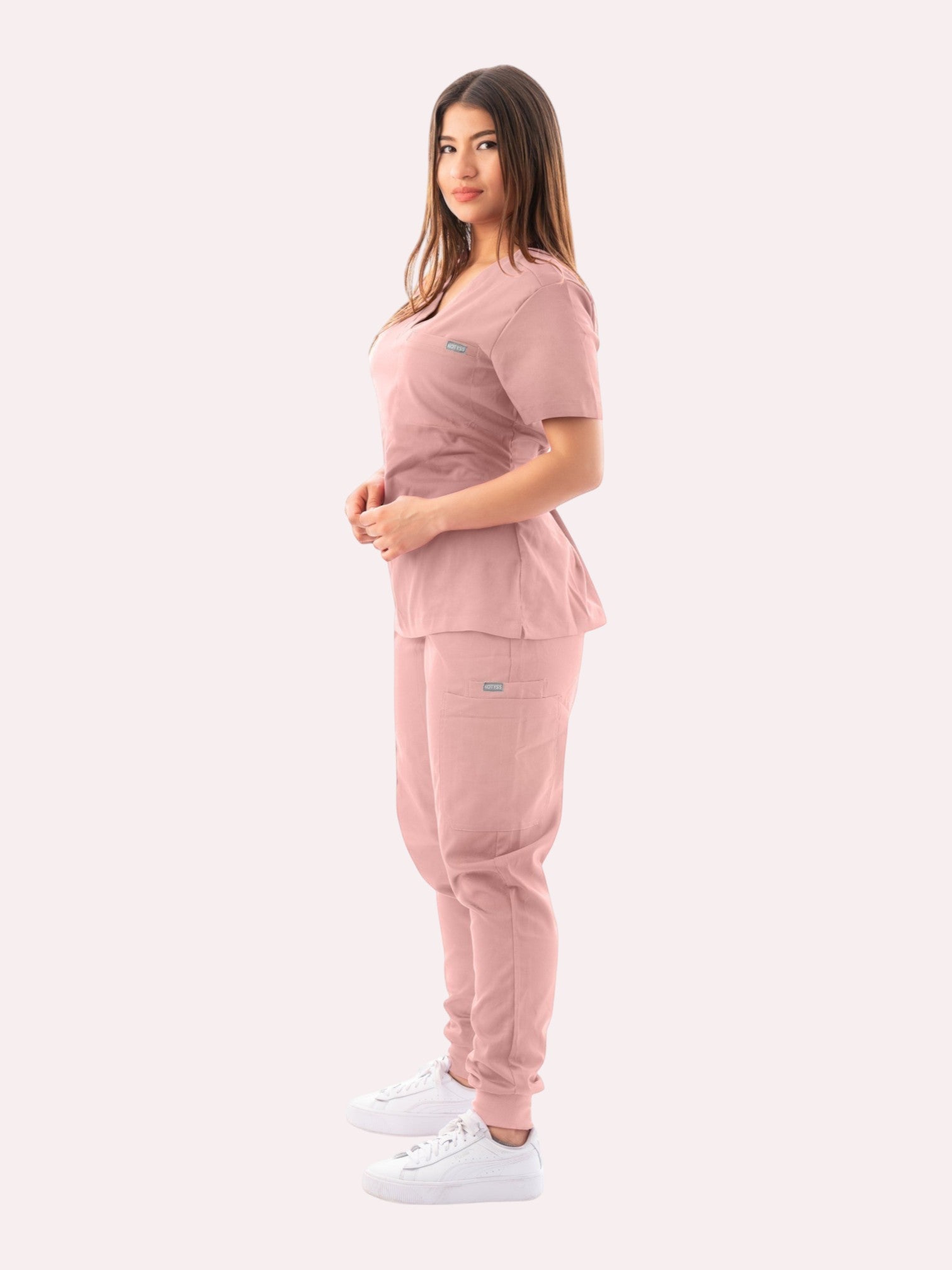 A woman wearing a dusty pink Kotyss V-neck scrub top and matching jogger pants with multiple pockets.
