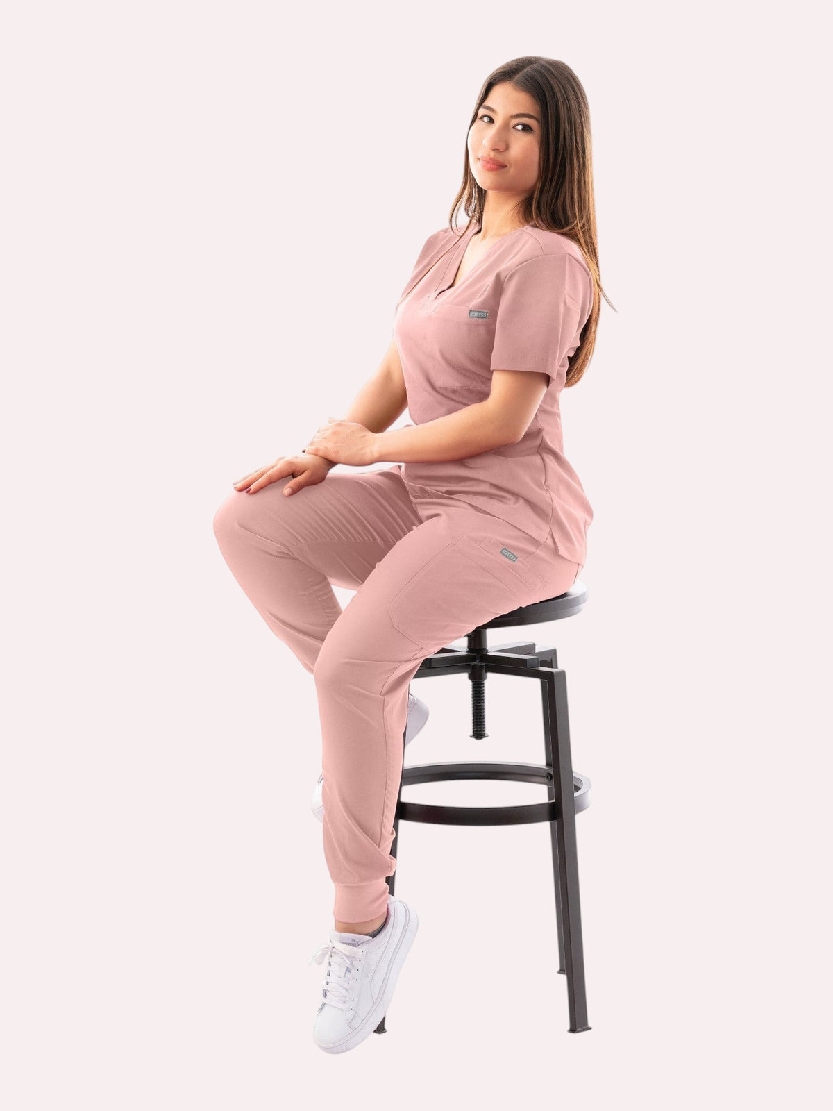 Woman wearing a dusty pink Kotyss V-neck scrub top and jogger pants set, sitting on a stool against a white background.