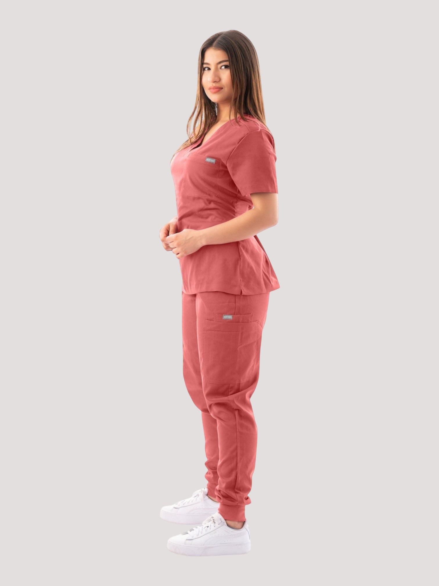 A woman wearing a Kotyss dusty rose V-neck scrub top and matching jogger pants with multiple utility pockets.