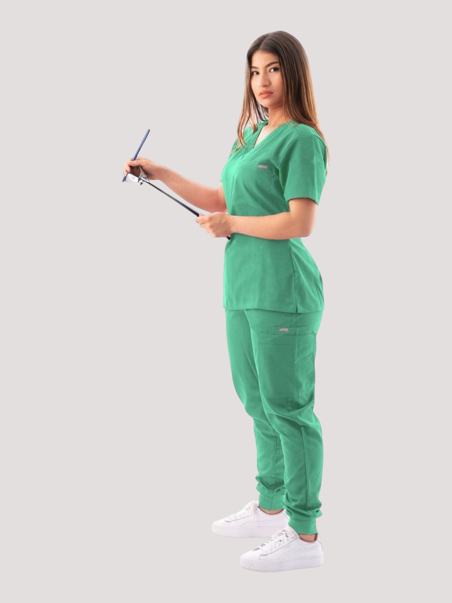 Kotyss green V-neck scrub top and jogger pants set worn by a woman holding a clipboard, side view.