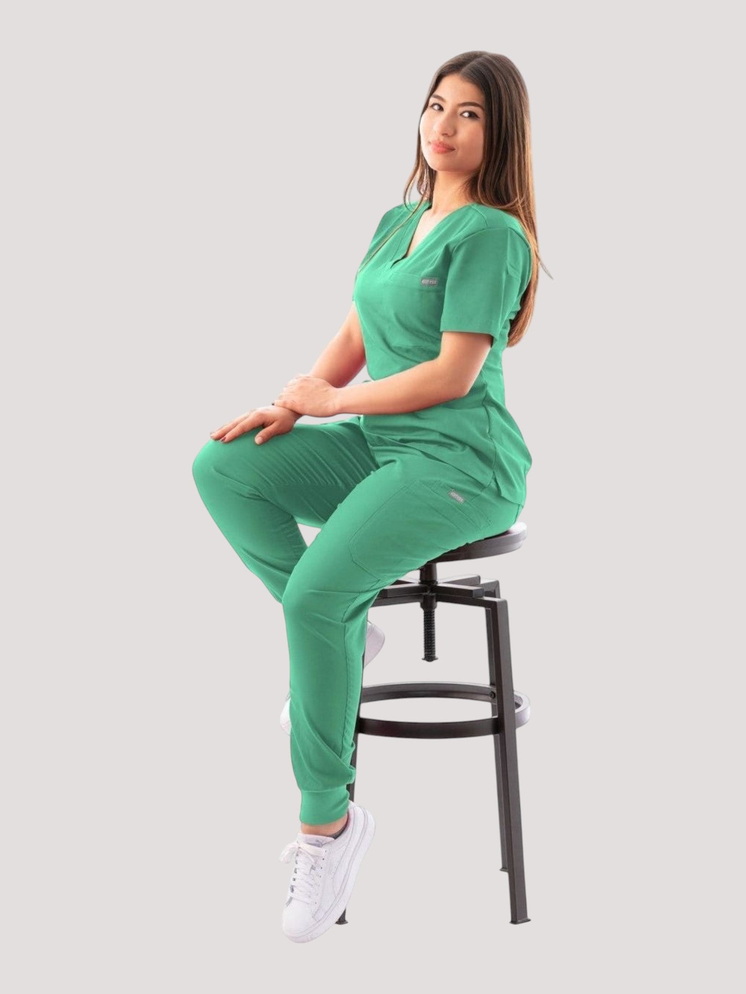Kotyss green V-neck scrub top and jogger pants set worn by a woman sitting on a stool, featuring 4-way stretch fabric.