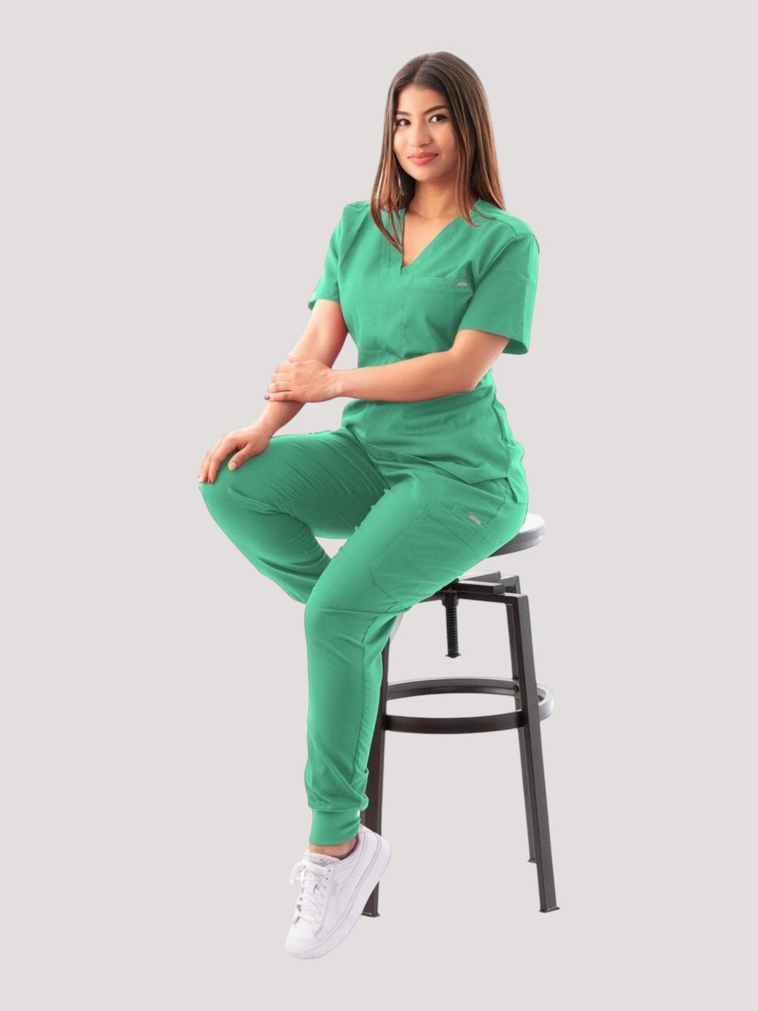 A woman wearing a green Kotyss V-neck scrub top and jogger pants set, sitting on a stool in a studio setting.