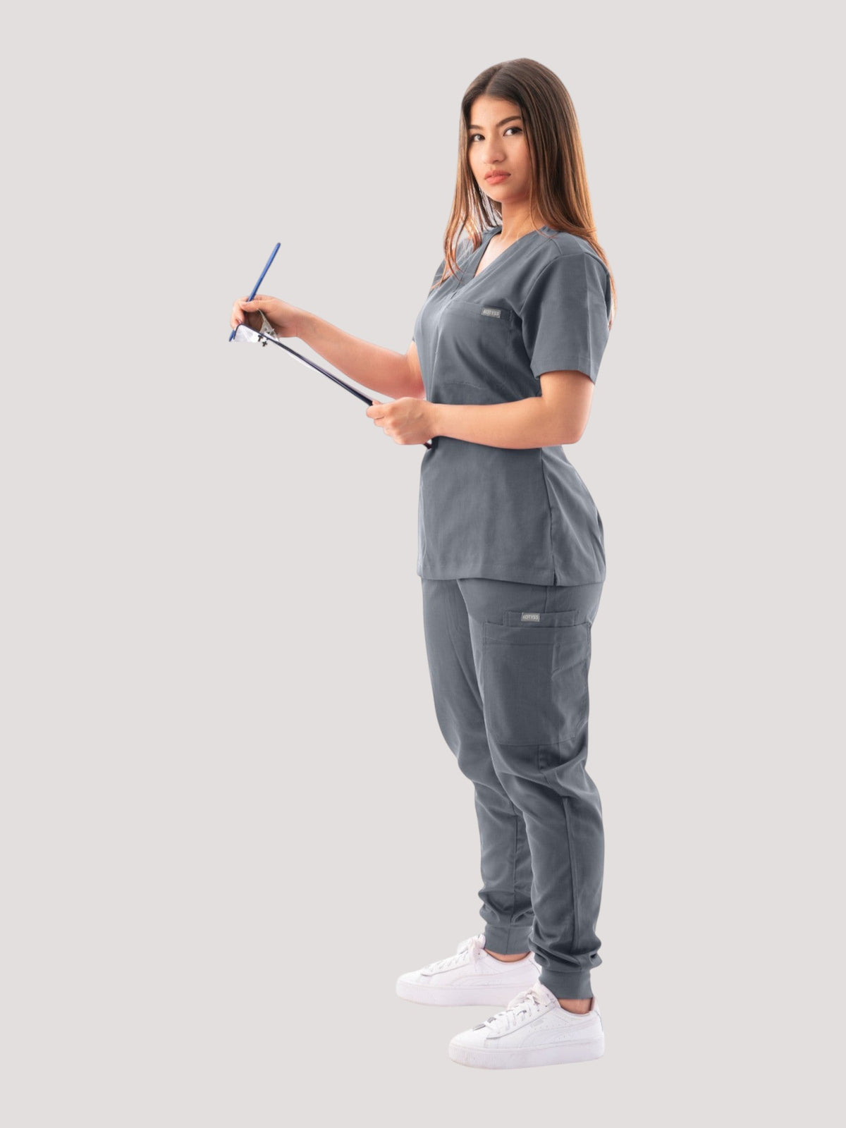 A woman wearing a grey Kotyss V-neck scrub top and matching jogger pants while holding a clipboard.