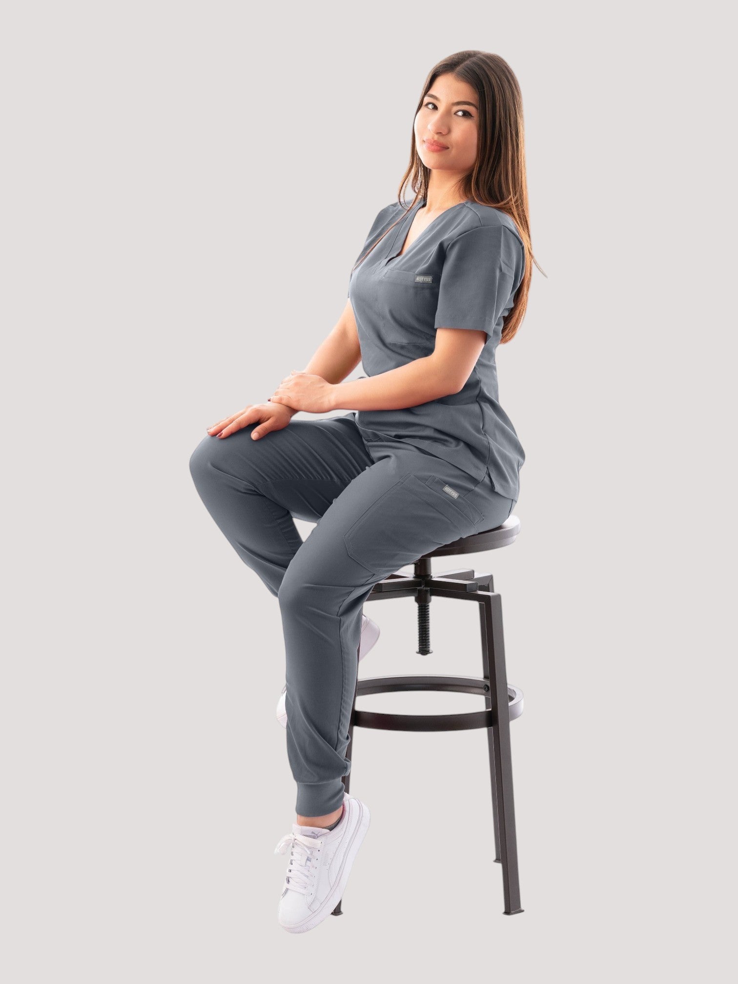A woman wearing a grey Kotyss V-neck scrub top and jogger pants set, sitting on a stool against a white background.