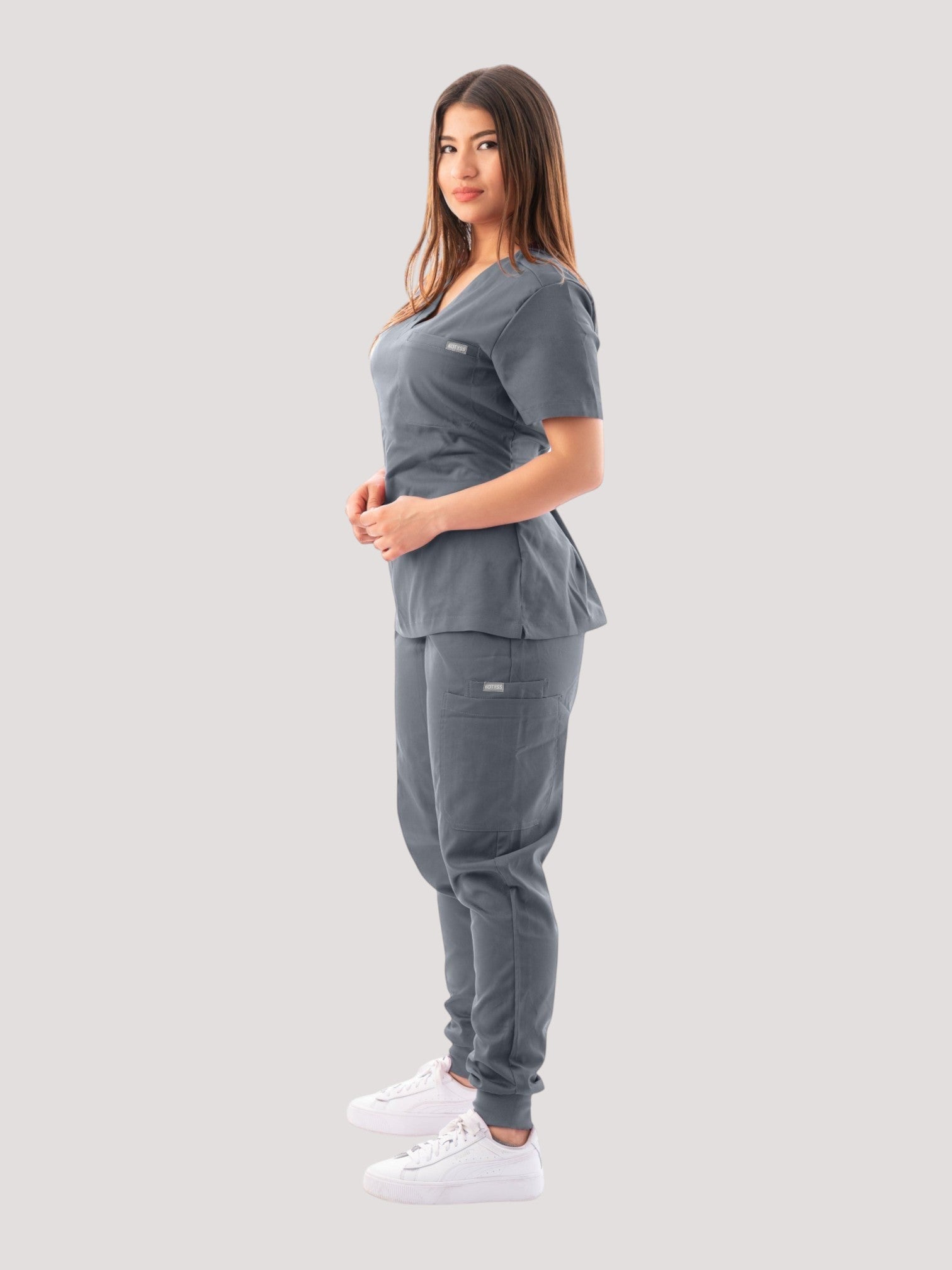 A woman wearing a grey Kotyss V-neck scrub top and matching jogger pants with multiple pockets on a white background.