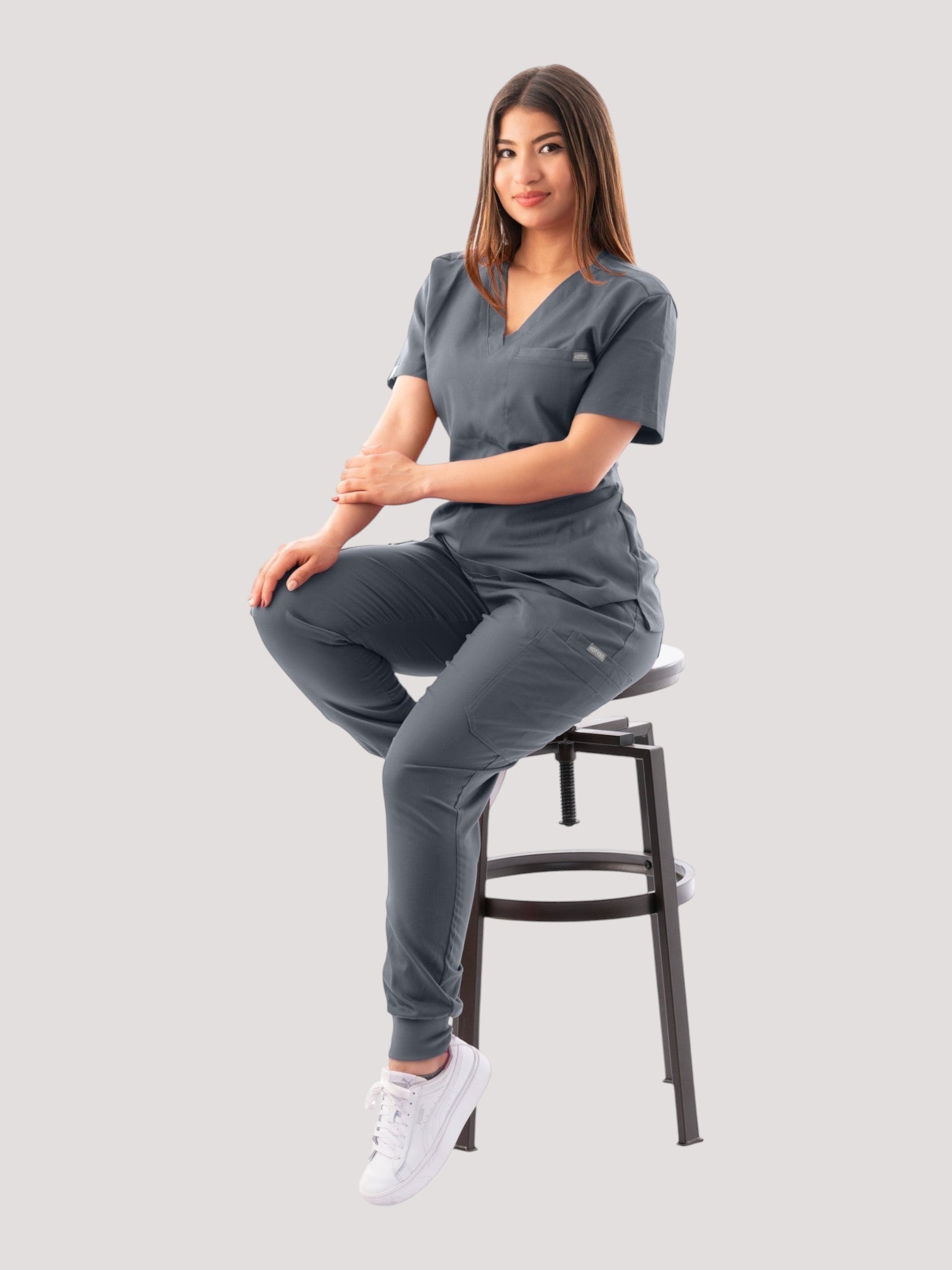 A woman wearing a grey Kotyss V-neck scrub top and matching jogger pants, sitting on a stool in a studio setting.