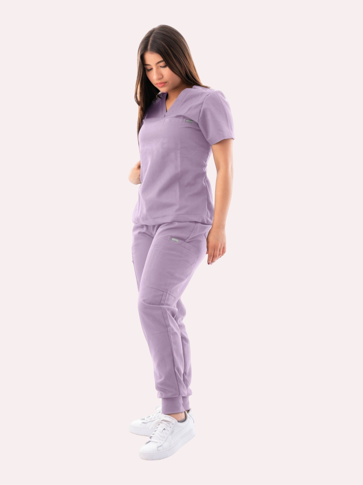 A woman wearing a Kotyss lavender V-neck scrub top and matching jogger pants with multiple utility pockets.