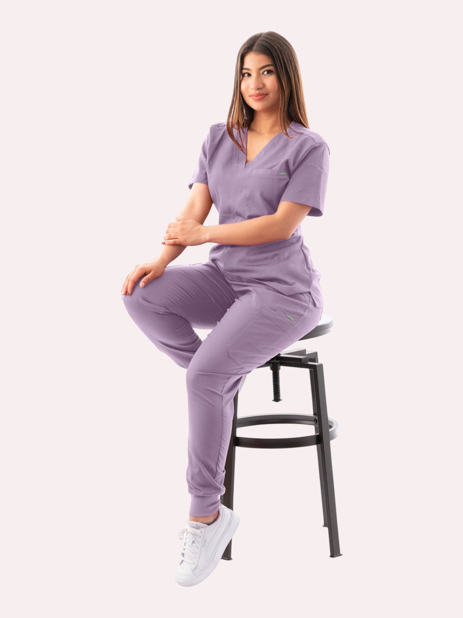 A woman wearing a lavender Kotyss V-neck scrub top and matching jogger pants while sitting on a stool.
