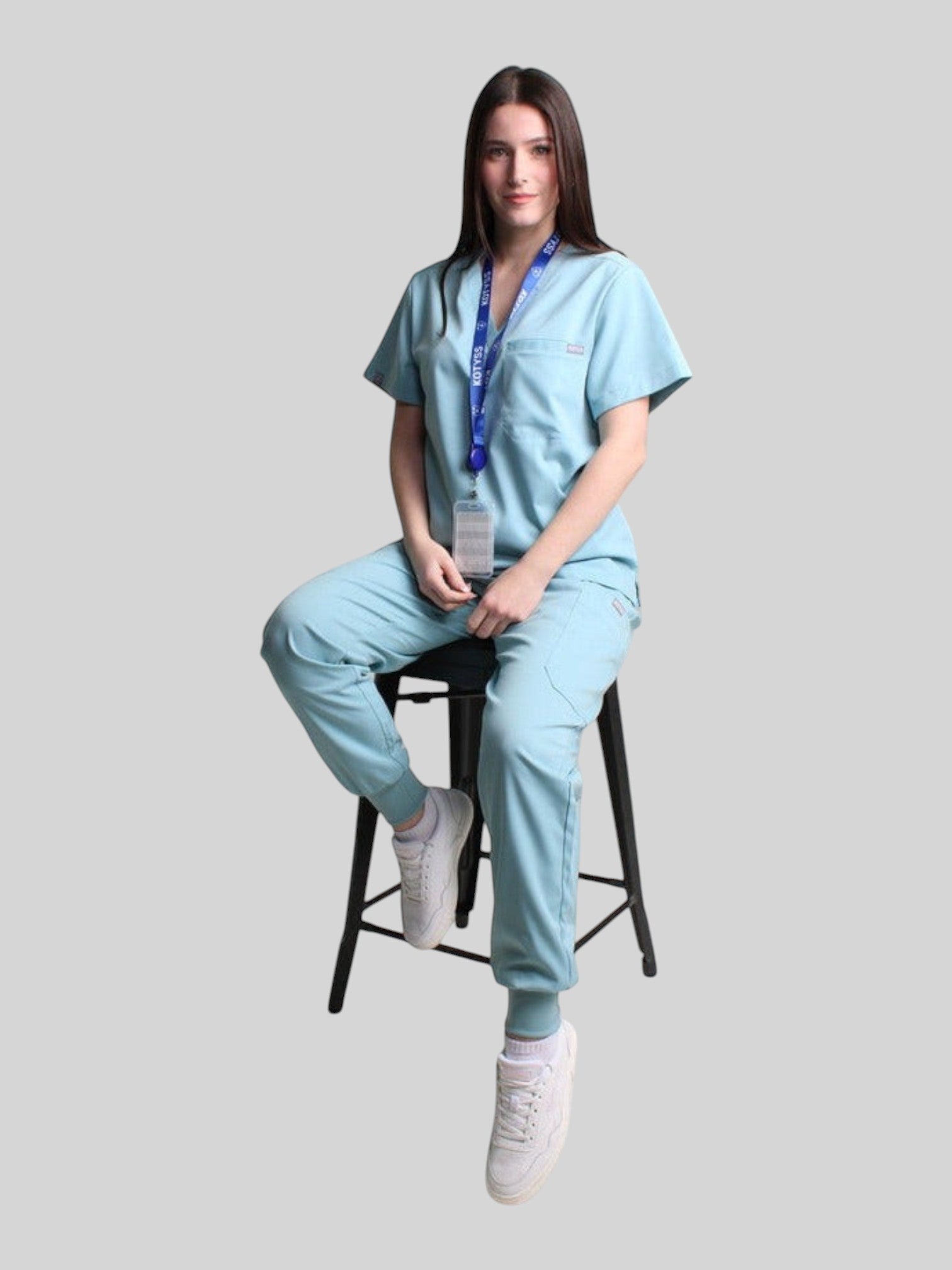 Woman wearing a light blue Kotyss V-neck scrub top and jogger pants set while sitting on a stool.