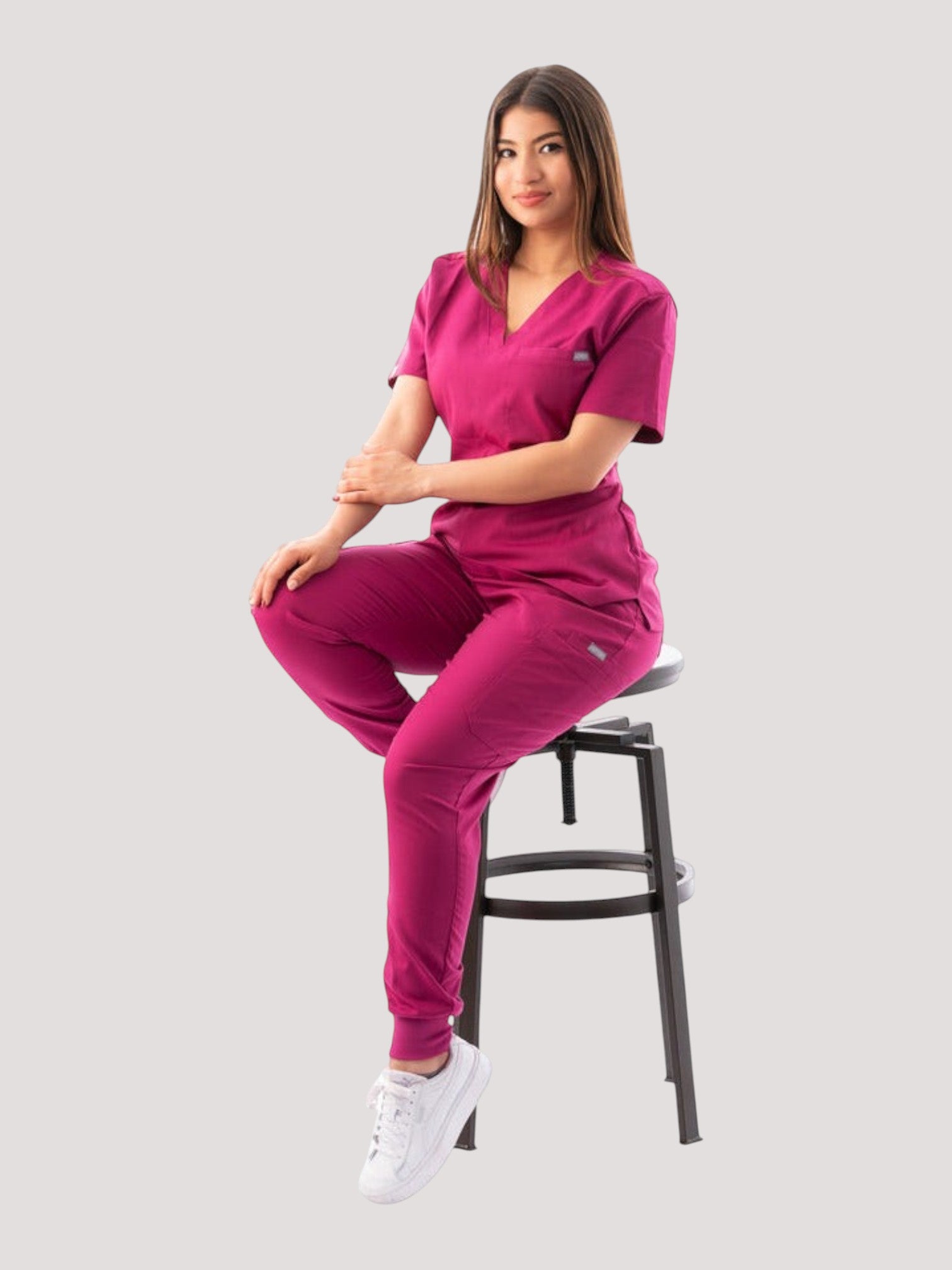 A woman wearing a magenta Kotyss V-neck scrub top and matching jogger pants while sitting on a stool.