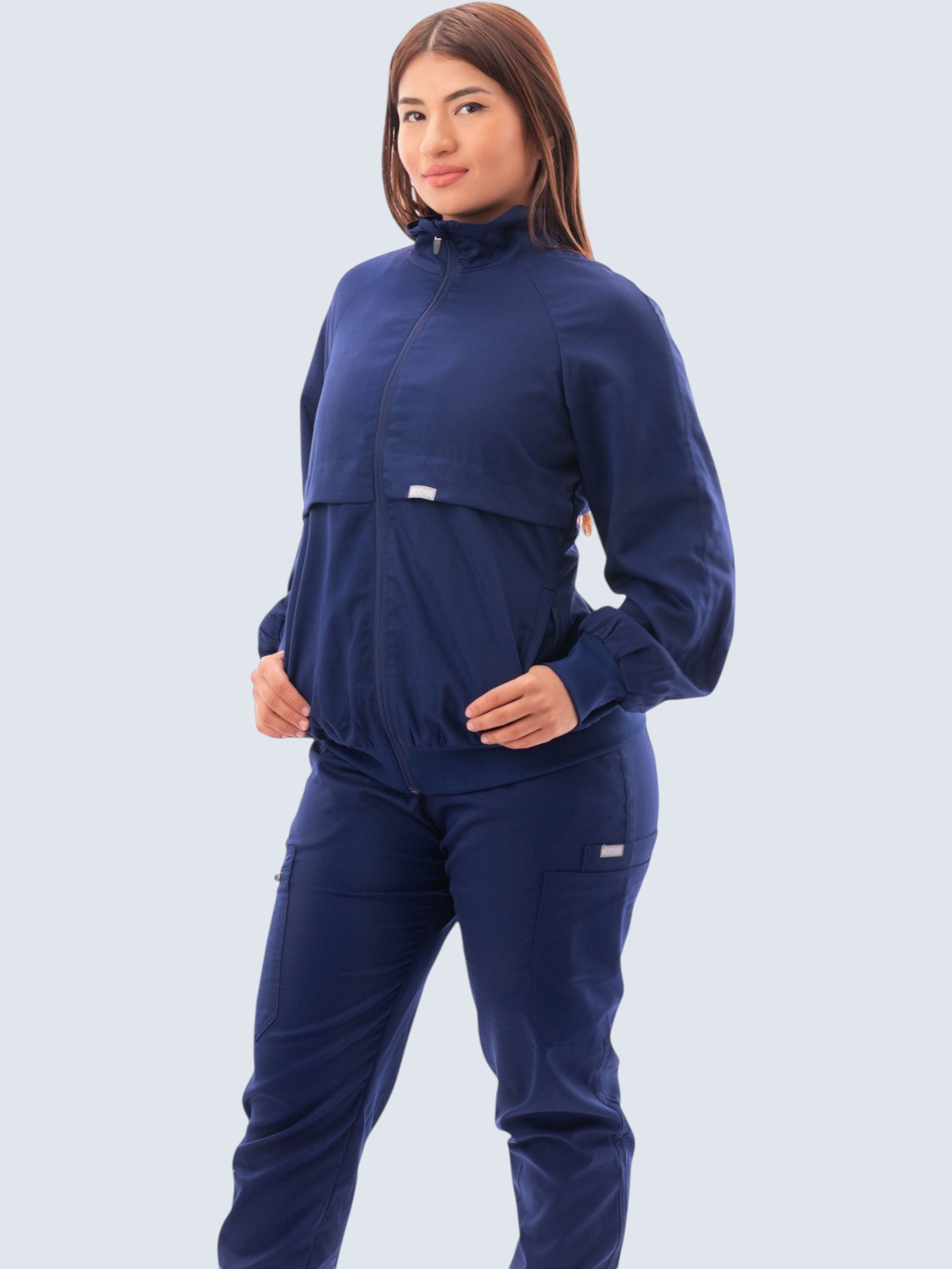 A model wearing a navy blue Kotyss women's scrub jacket with a high windbreaker neck and ribbed knit cuffs.