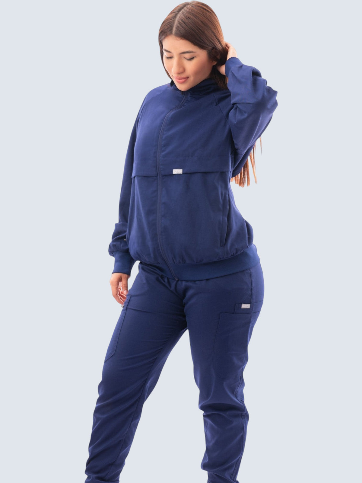 A woman wearing a navy blue Kotyss scrub jacket with a high neck, zip front, and ribbed cuffs over matching scrubs.