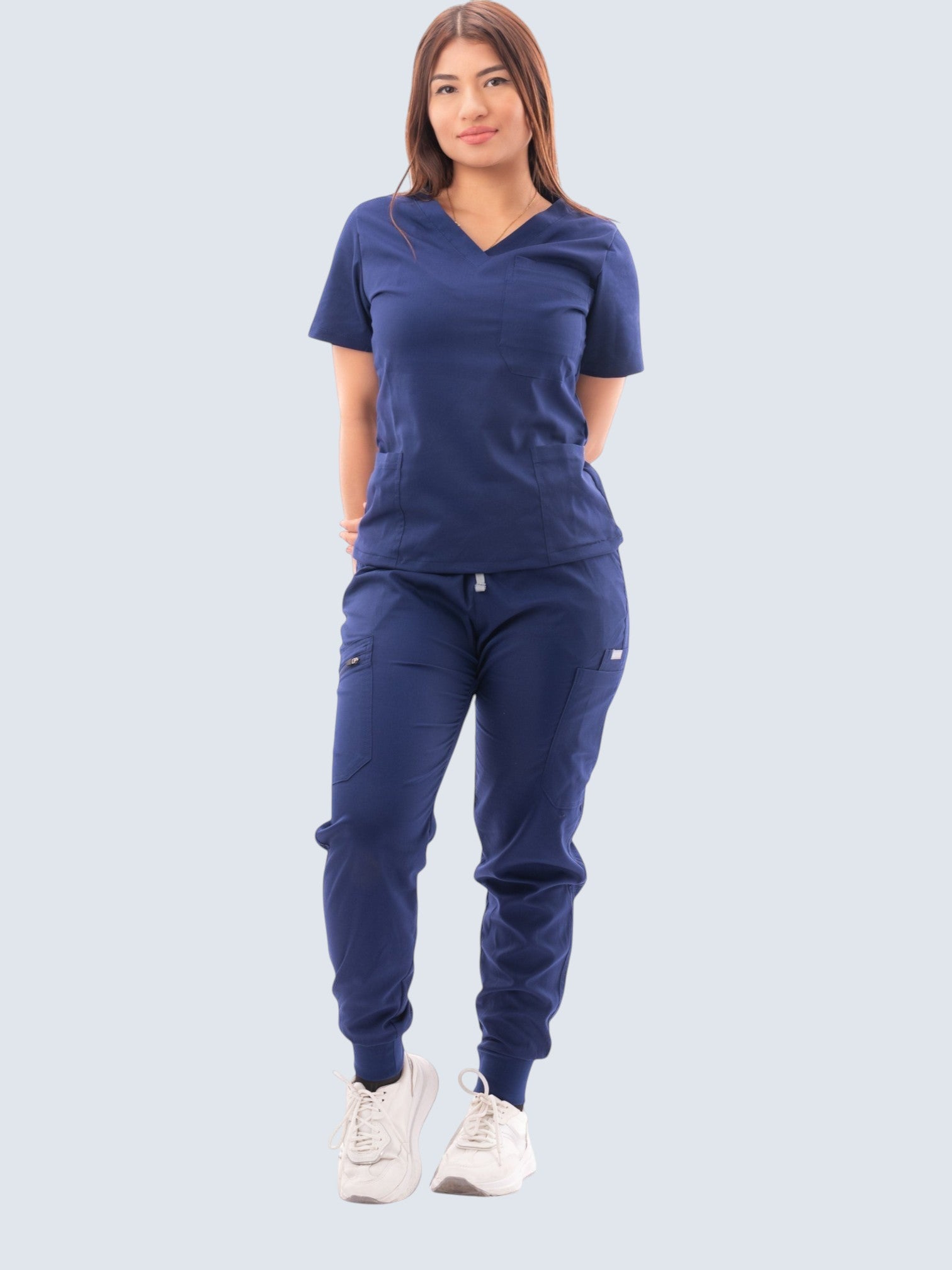 A woman wearing a Kotyss navy blue V-neck scrub top and matching multi-pocket jogger pants with white sneakers.