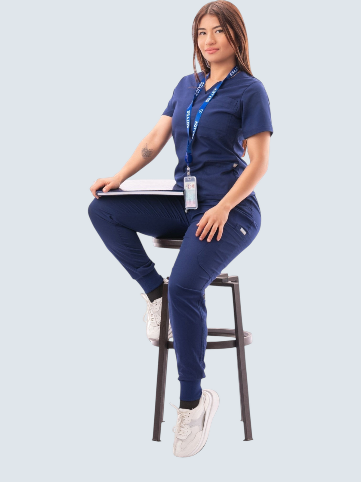 Woman wearing a navy blue Kotyss V-neck scrub top and jogger pants set with multiple pockets, sitting on a stool.