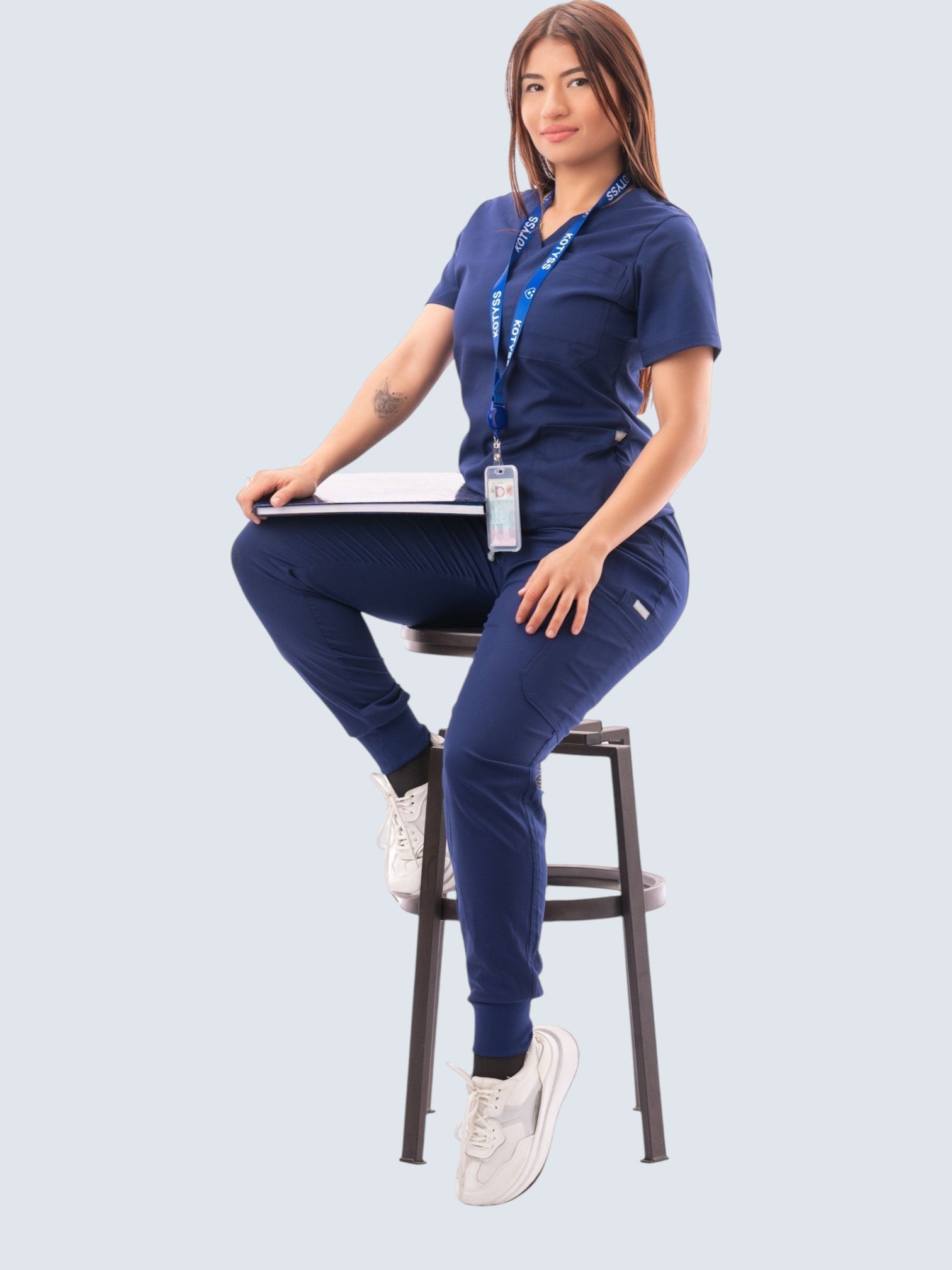 Kotyss navy blue V-neck scrub top and jogger pants set worn by a woman sitting on a stool with a lanyard