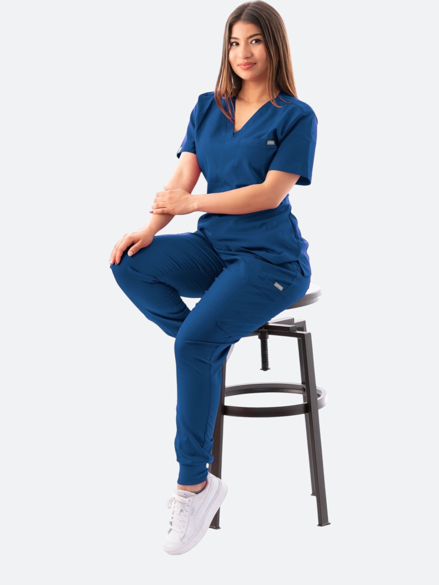 A woman wearing a navy blue Kotyss V-neck scrub top and jogger pants set, sitting on a stool.
