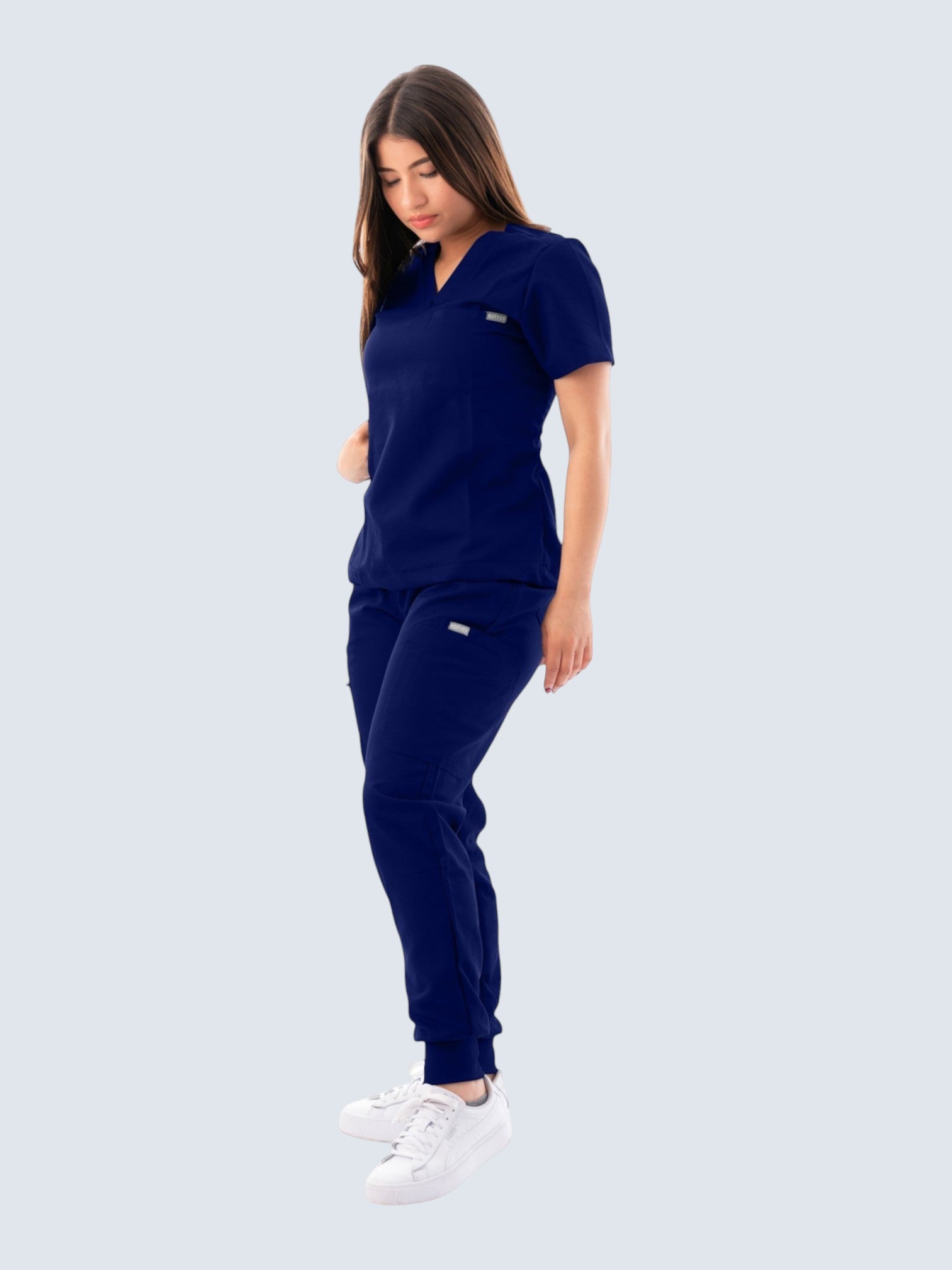 A woman wearing a navy blue Kotyss V-neck scrub top and matching jogger pants with multiple pockets.