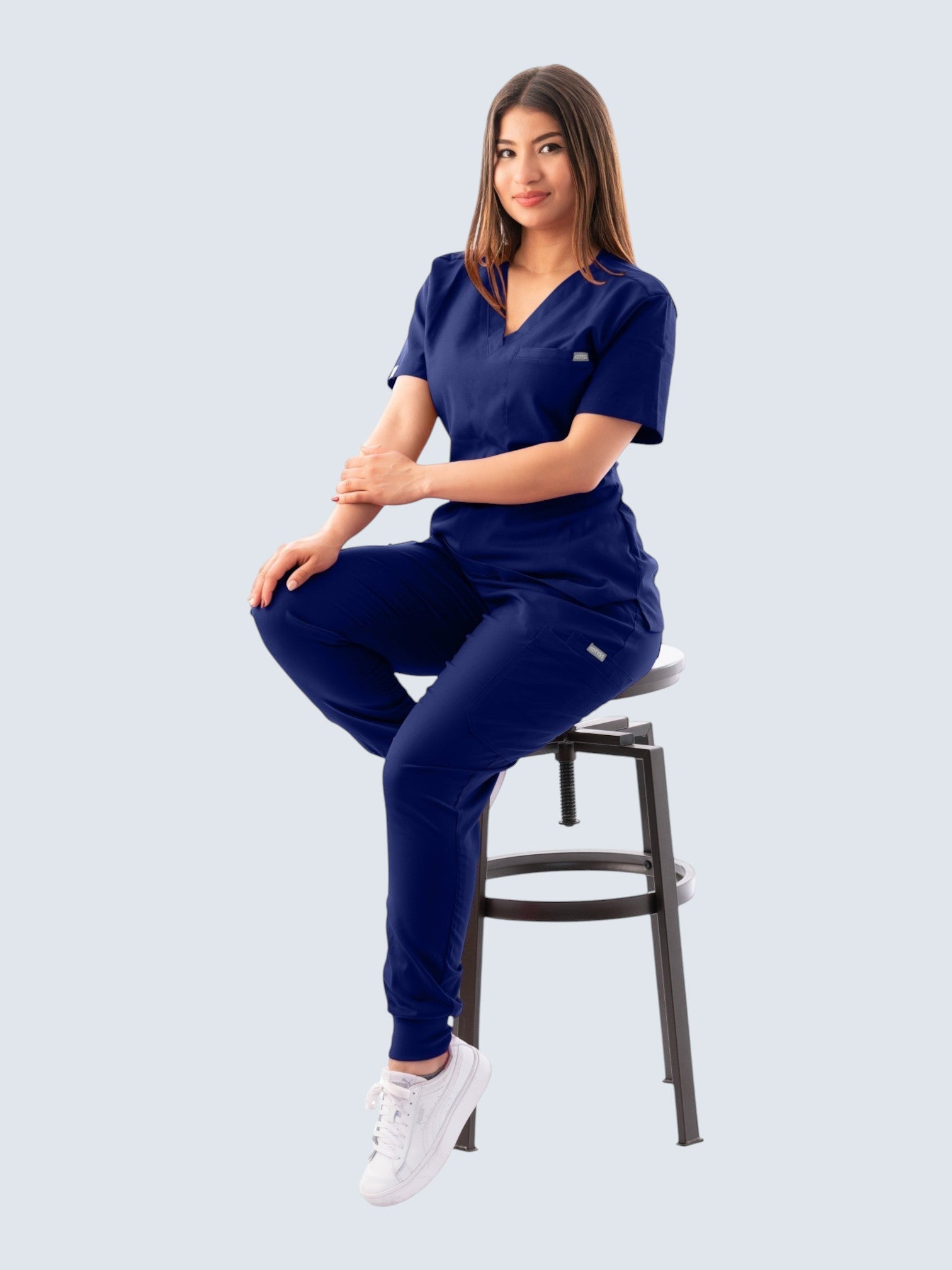 Kotyss navy blue V-neck scrub top and jogger pants set worn by a woman sitting on a stool.