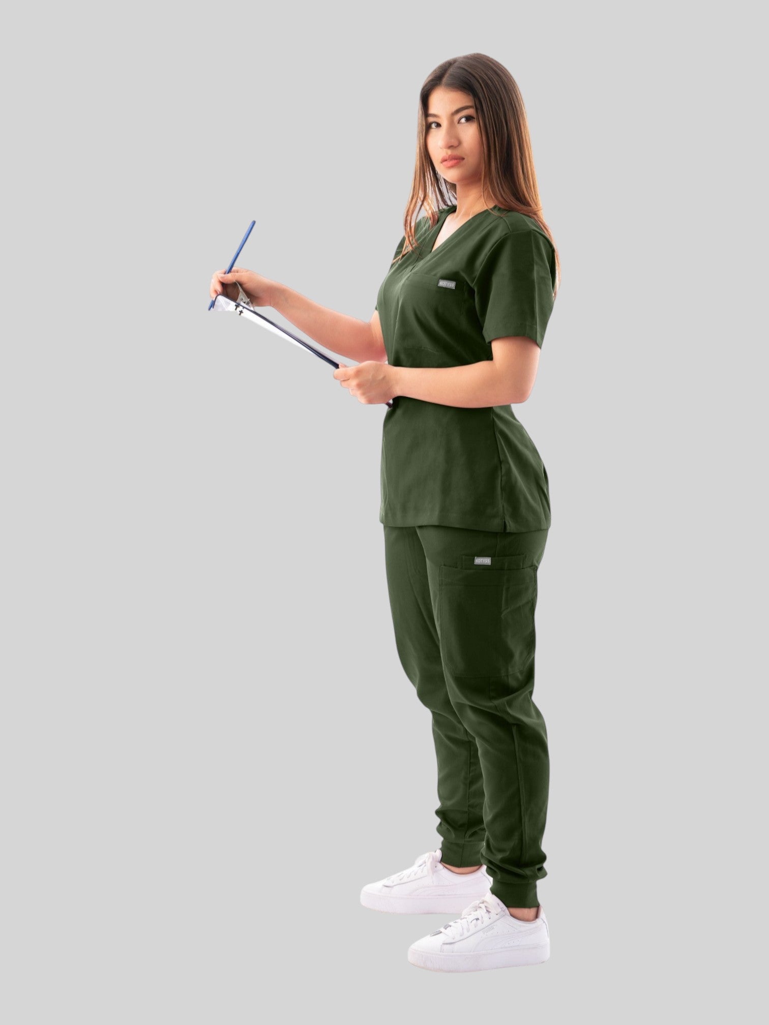 Kotyss olive green V-neck scrub top and jogger pants set worn by a woman holding a clipboard