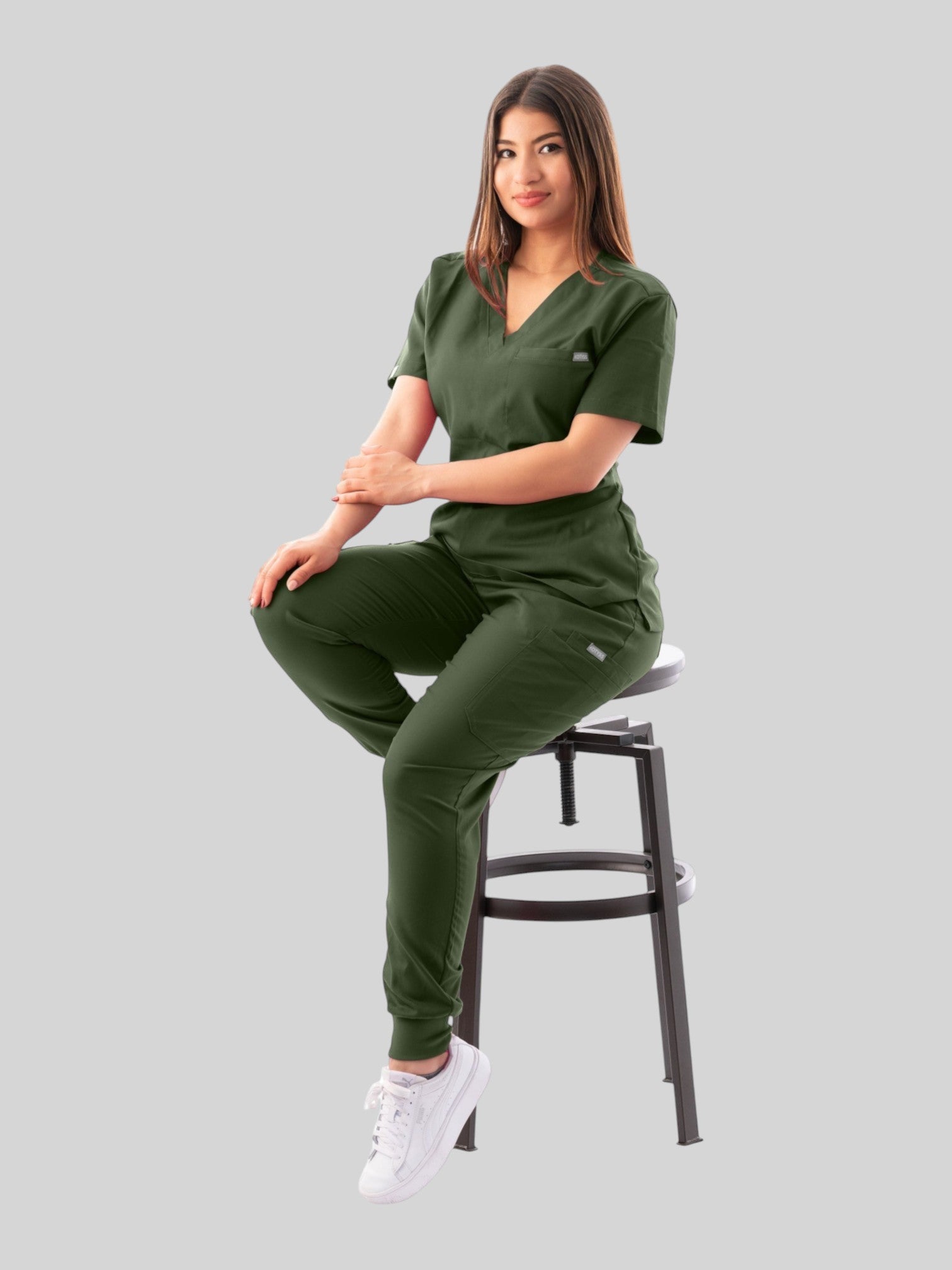 Kotyss olive green V-neck scrub top and jogger pants set worn by a woman sitting on a stool