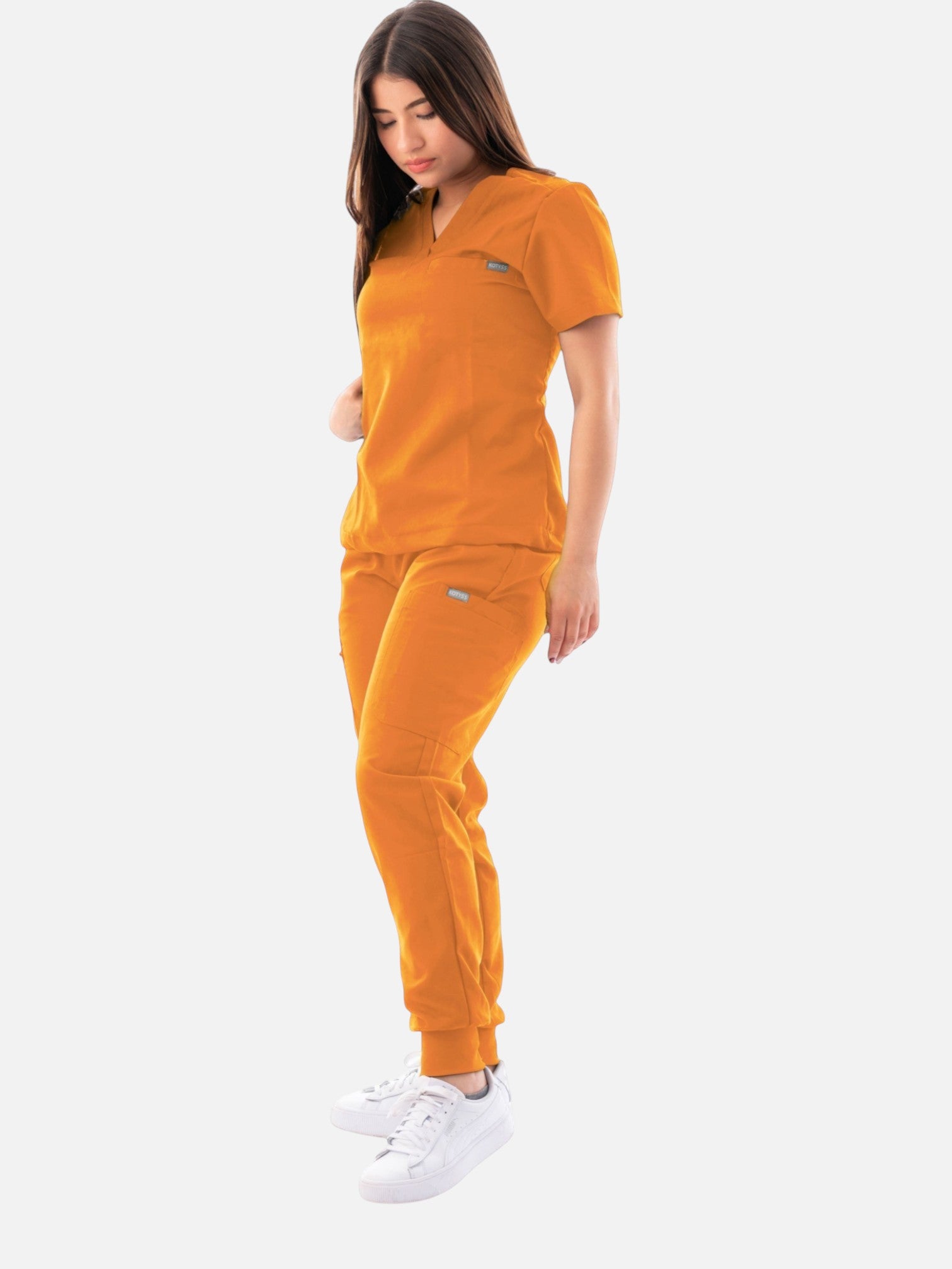 A woman wearing a vibrant orange Kotyss V-neck scrub top and matching jogger pants with white sneakers.