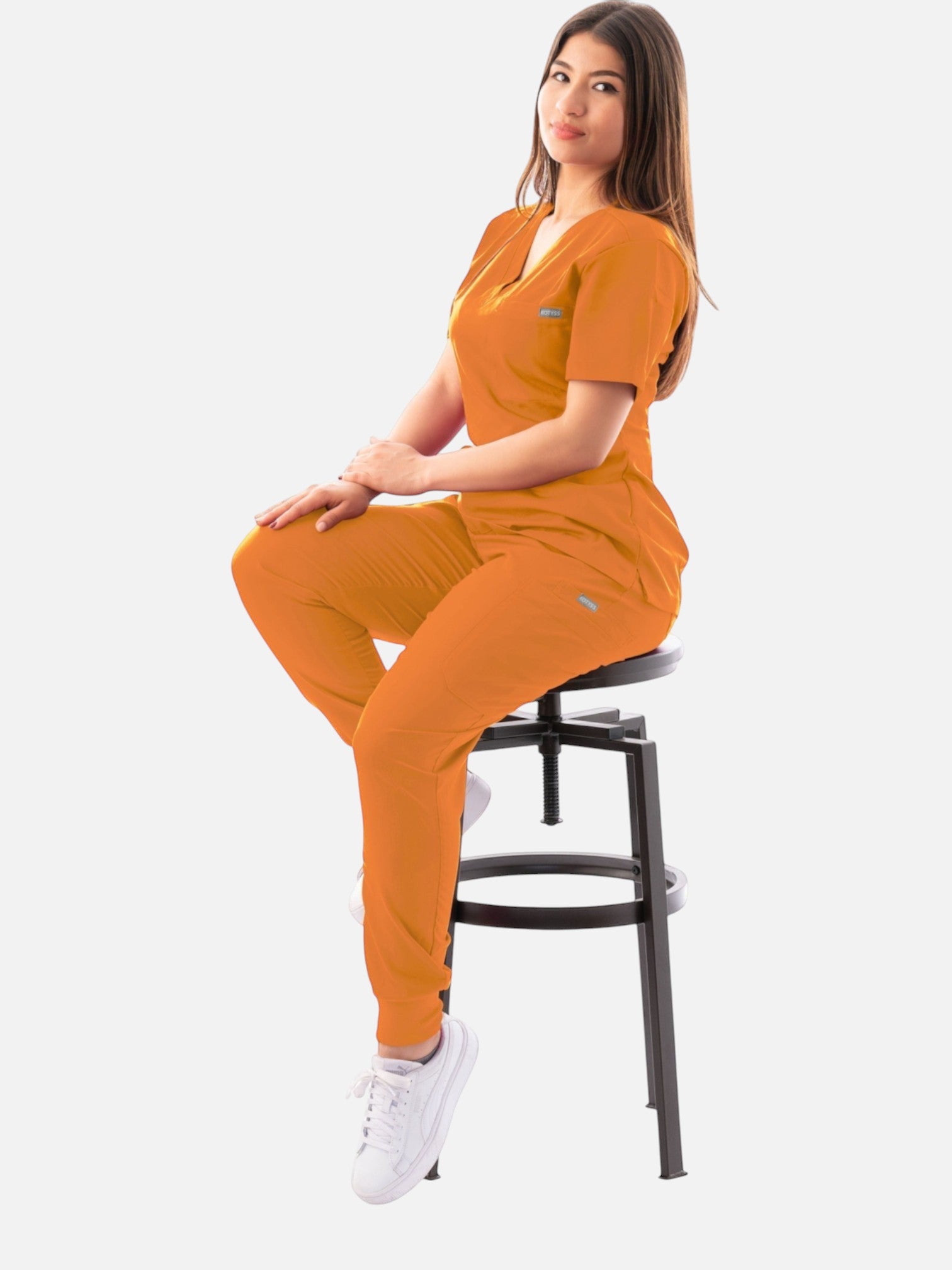 A woman wearing a vibrant orange Kotyss V-neck scrub top and matching jogger pants while sitting on a stool.