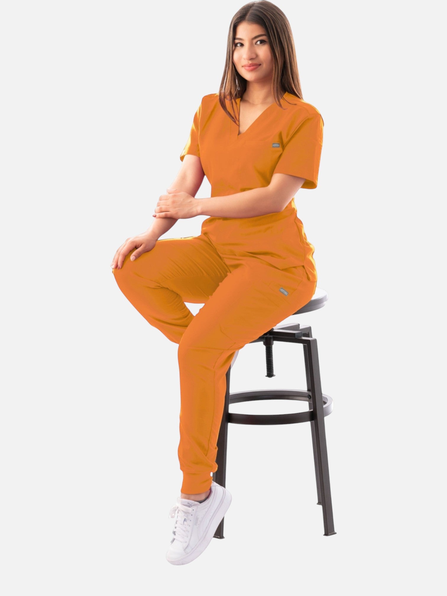 A woman wearing a vibrant orange Kotyss V-neck scrub top and matching jogger pants while sitting on a stool.