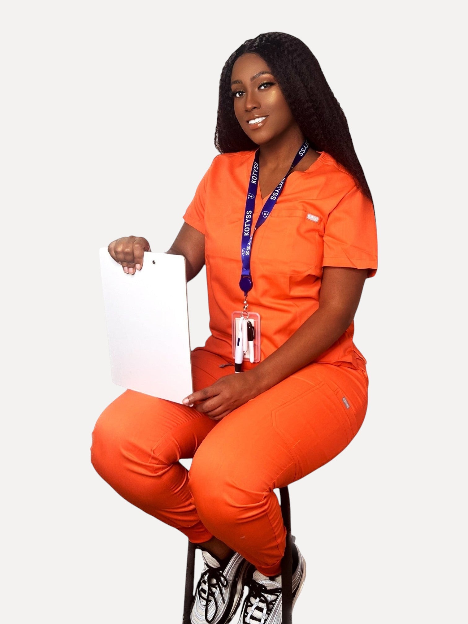 Kotyss model wearing an orange V-neck scrub top and jogger pants set with a clipboard, isolated on white background.