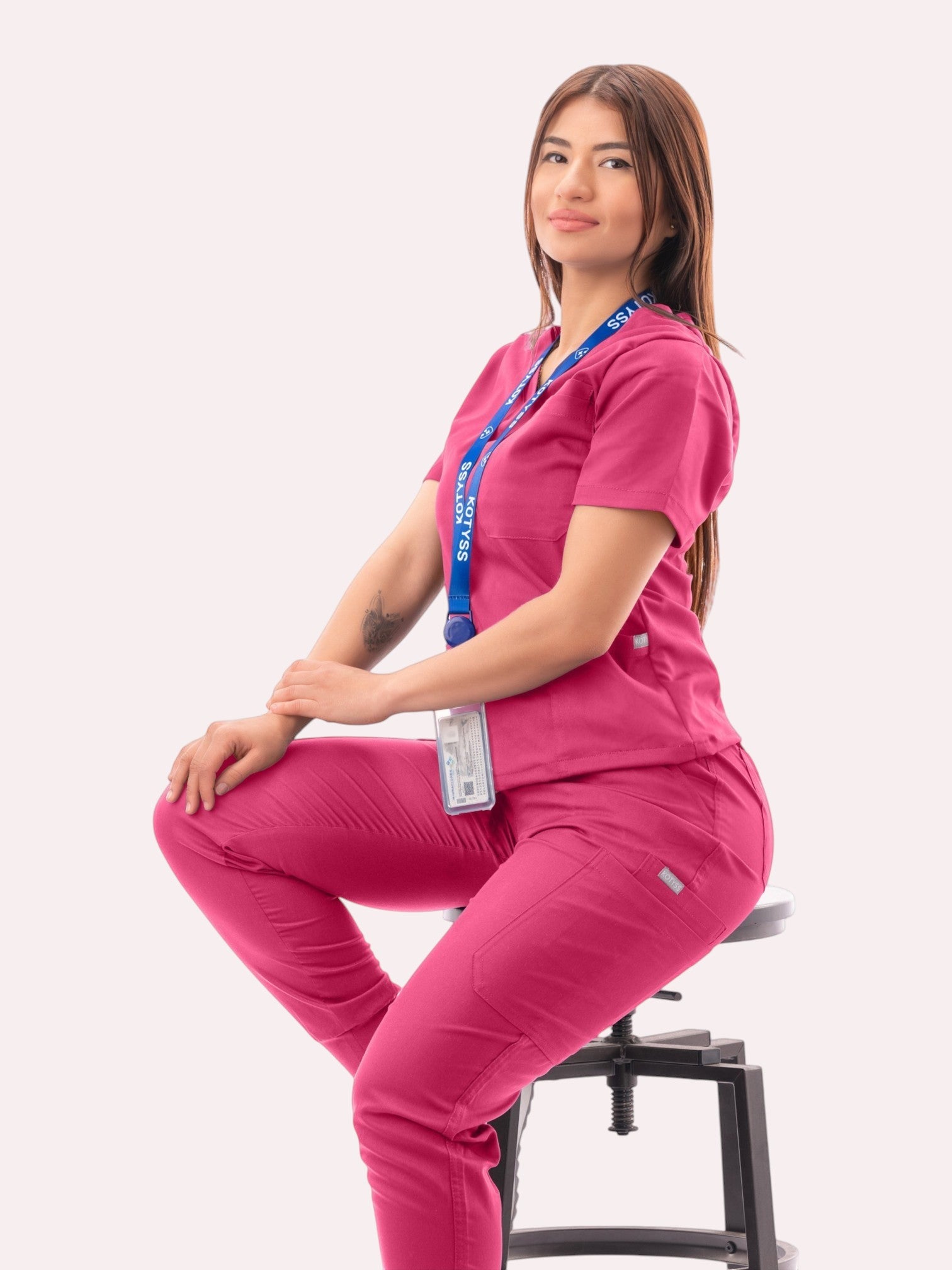 A woman wearing a vibrant pink Kotyss V-neck scrub top and matching jogger pants while sitting on a stool.