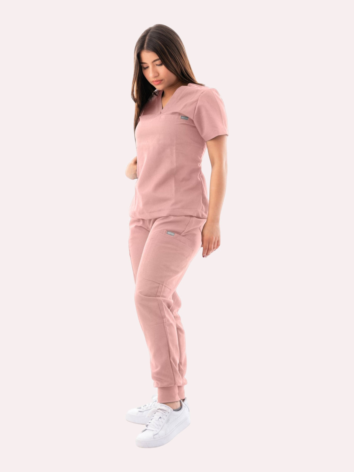Model wearing a dusty pink Kotyss V-neck scrub top and matching jogger pants with multiple utility pockets.