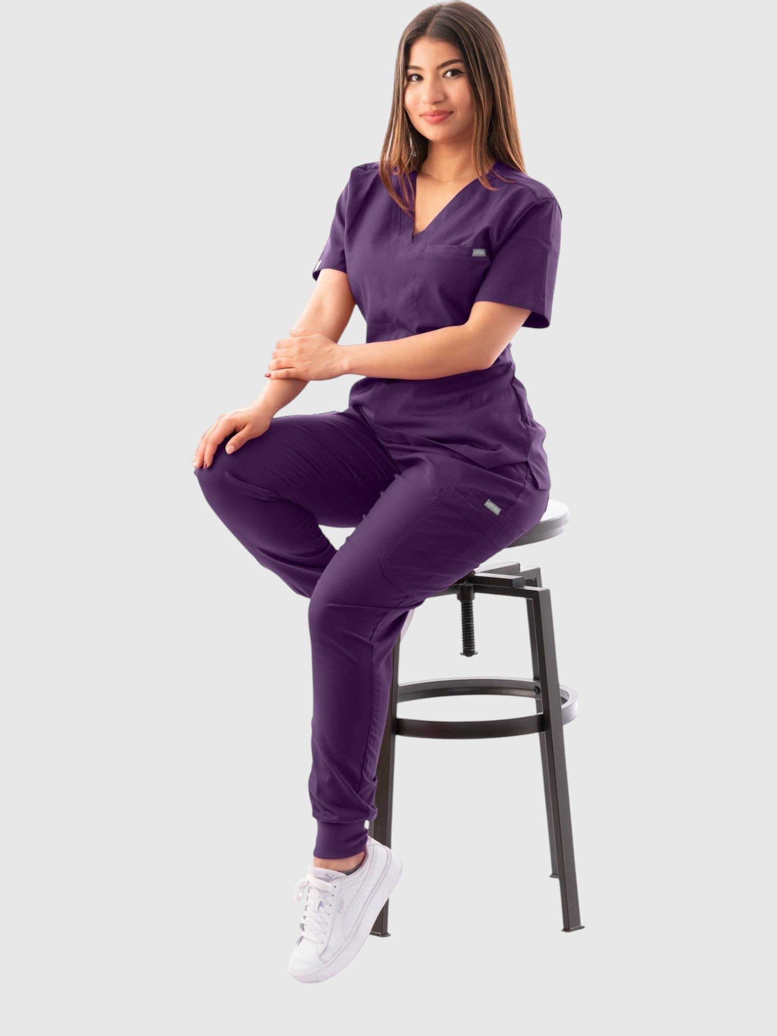 A woman wearing a purple Kotyss V-neck scrub top and jogger pants set, sitting on a stool in a studio setting.