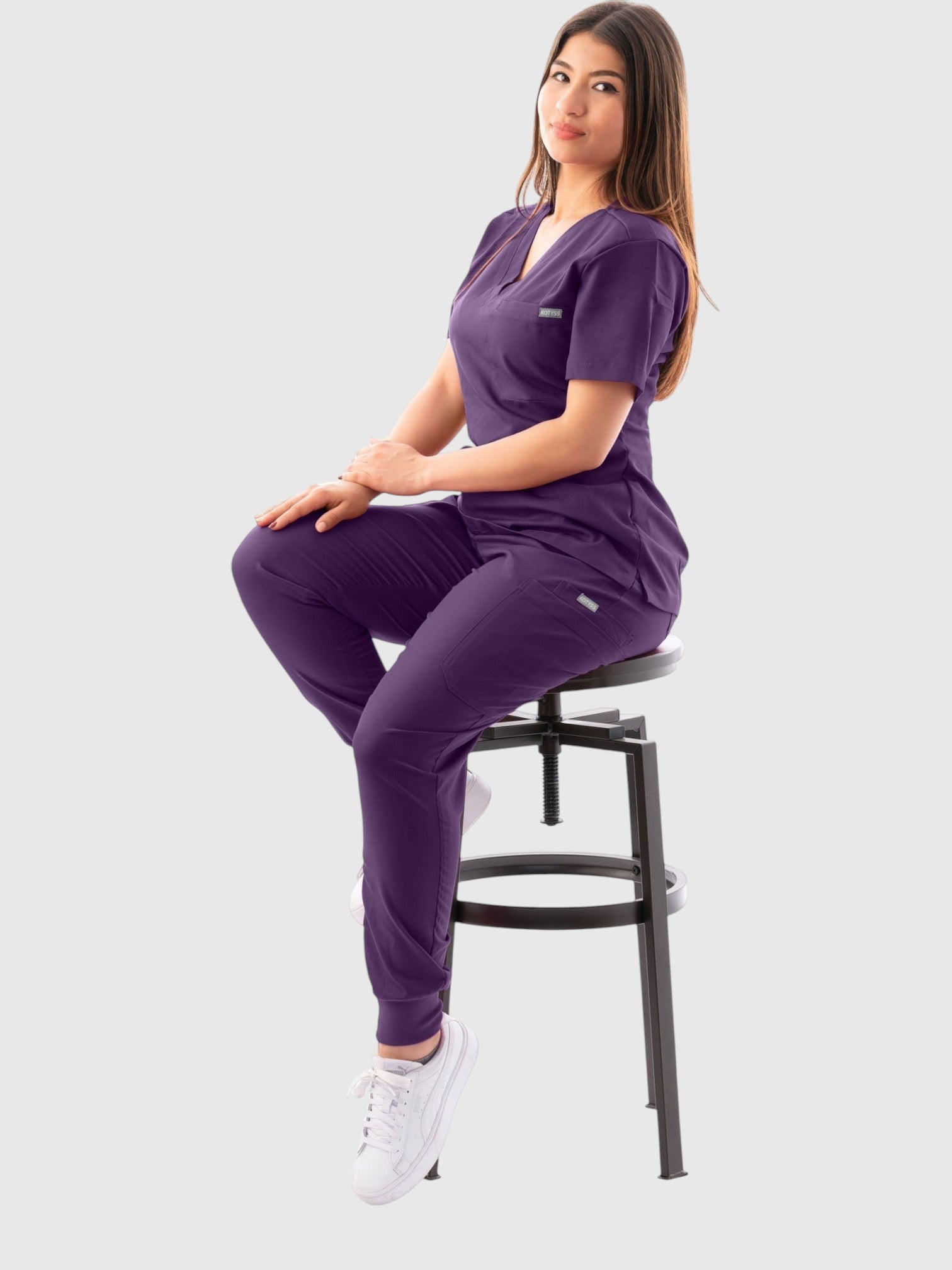 Kotyss women's purple V-neck scrub top and jogger pants set worn by a model sitting on a stool